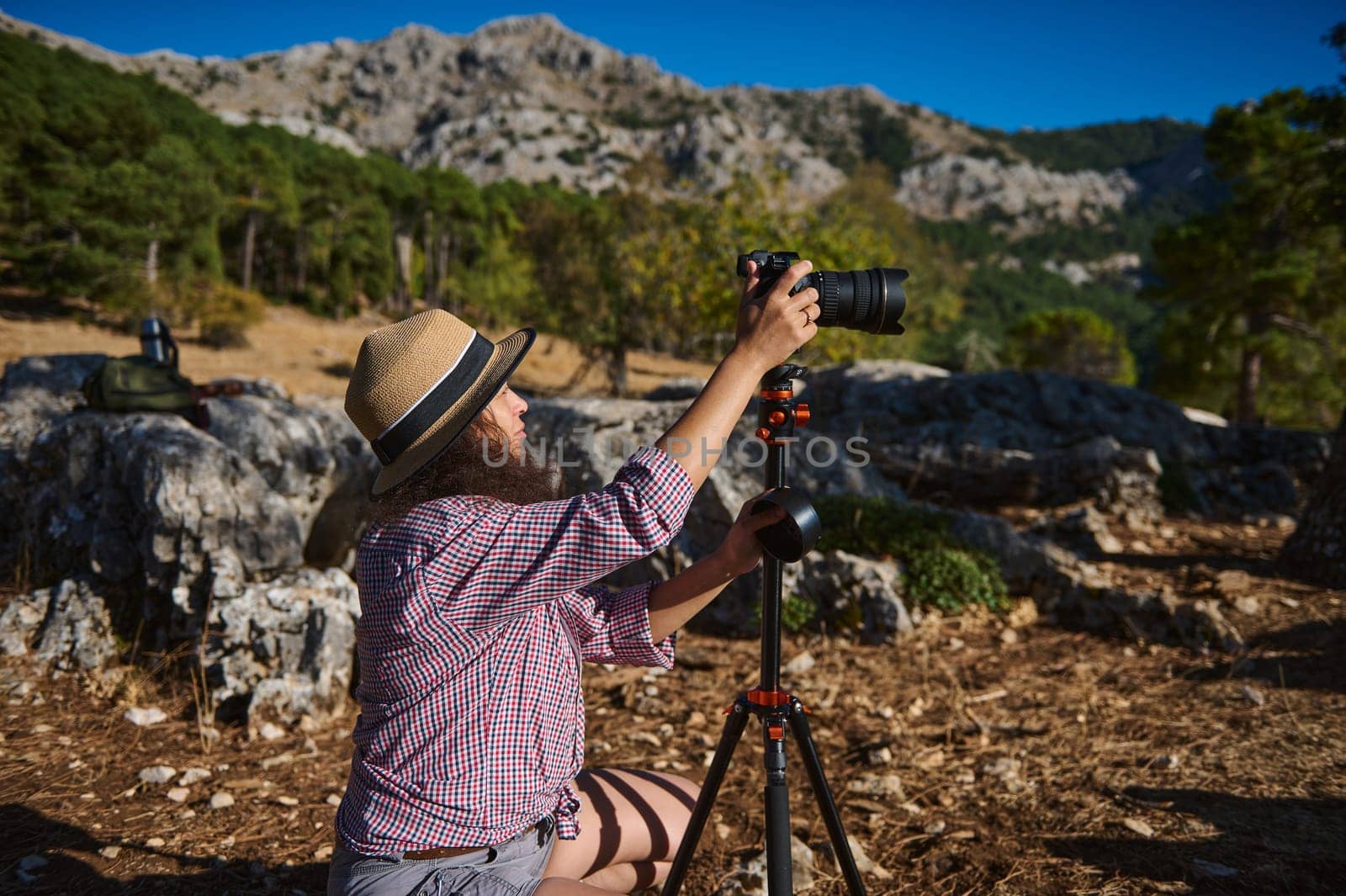 A woman in a hat and casual outfit prepares her camera on a tripod amidst a rugged mountain setting, capturing the essence of outdoor photography and exploration.