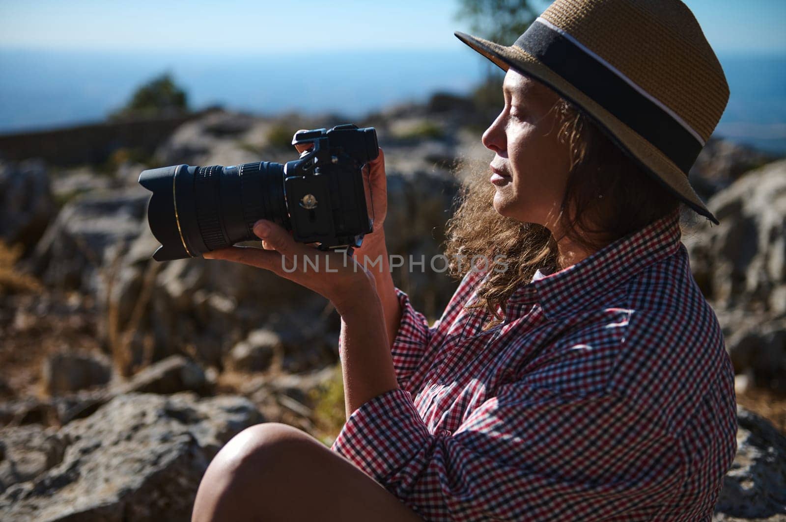A woman, wearing a straw hat and plaid shirt, sits outdoors holding a camera. She is enjoying nature while photographing the rocky landscape. A sense of adventure and exploration.