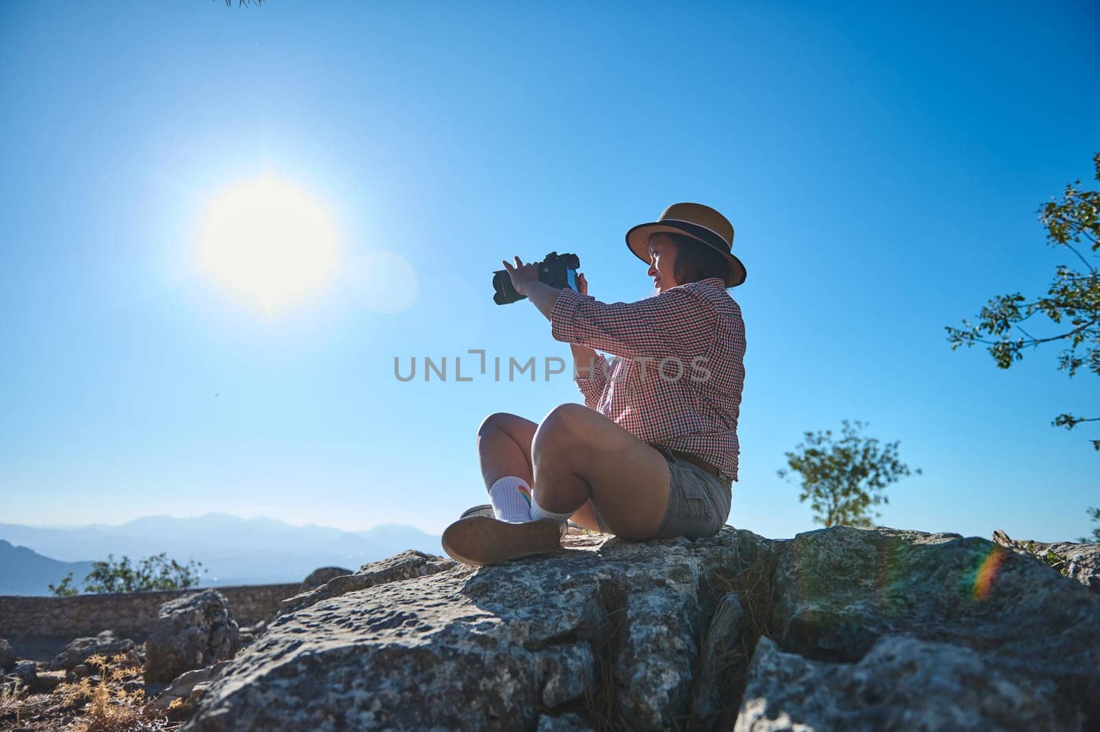 A woman sits on rocky terrain, using binoculars under a clear blue sky. The scene reflects adventure, exploration, and appreciation of nature's beauty.