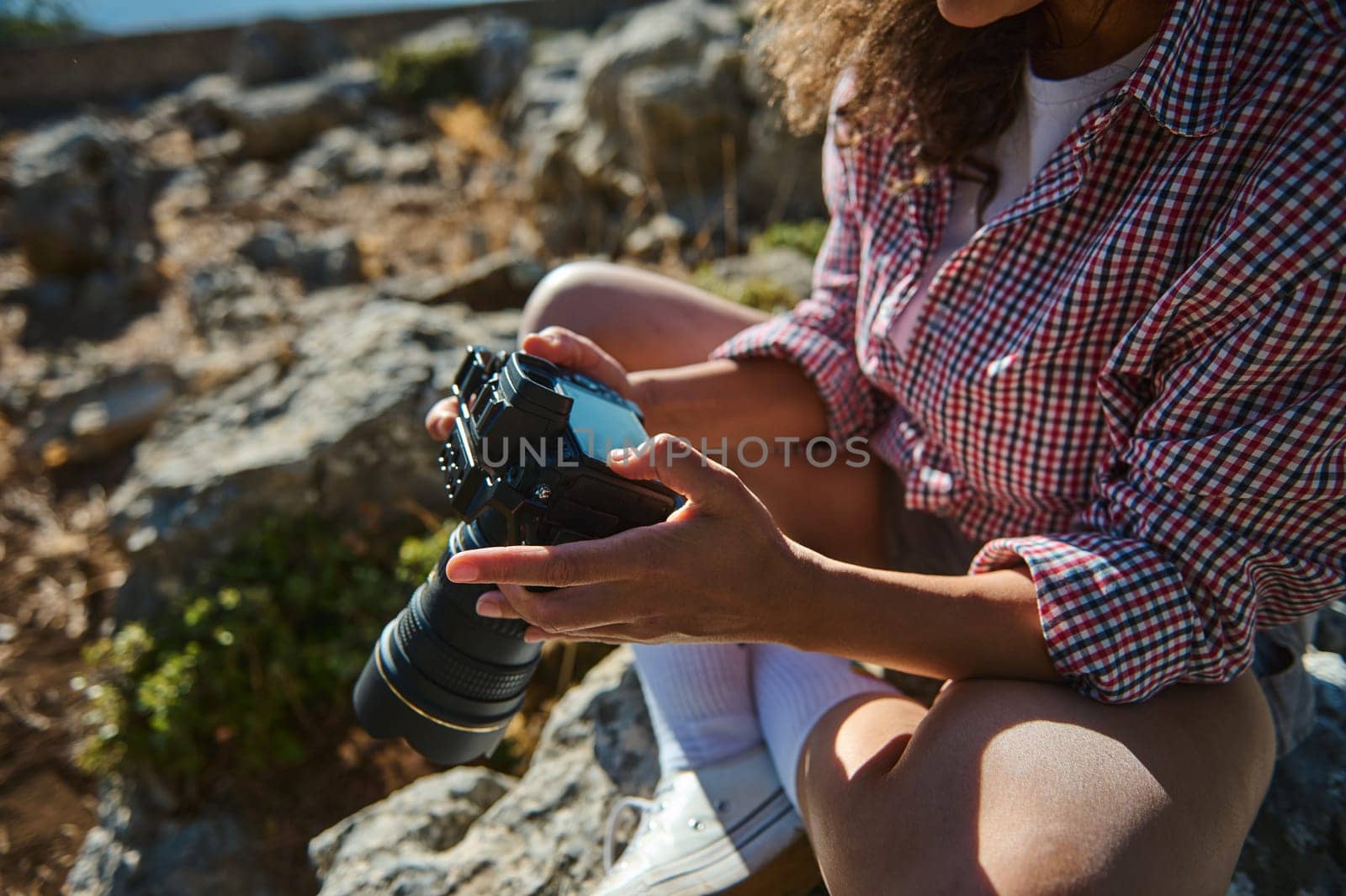 A woman in a plaid shirt sits on rocky terrain, adjusting her camera settings. The scene captures exploration, photography, and leisure in a natural environment.