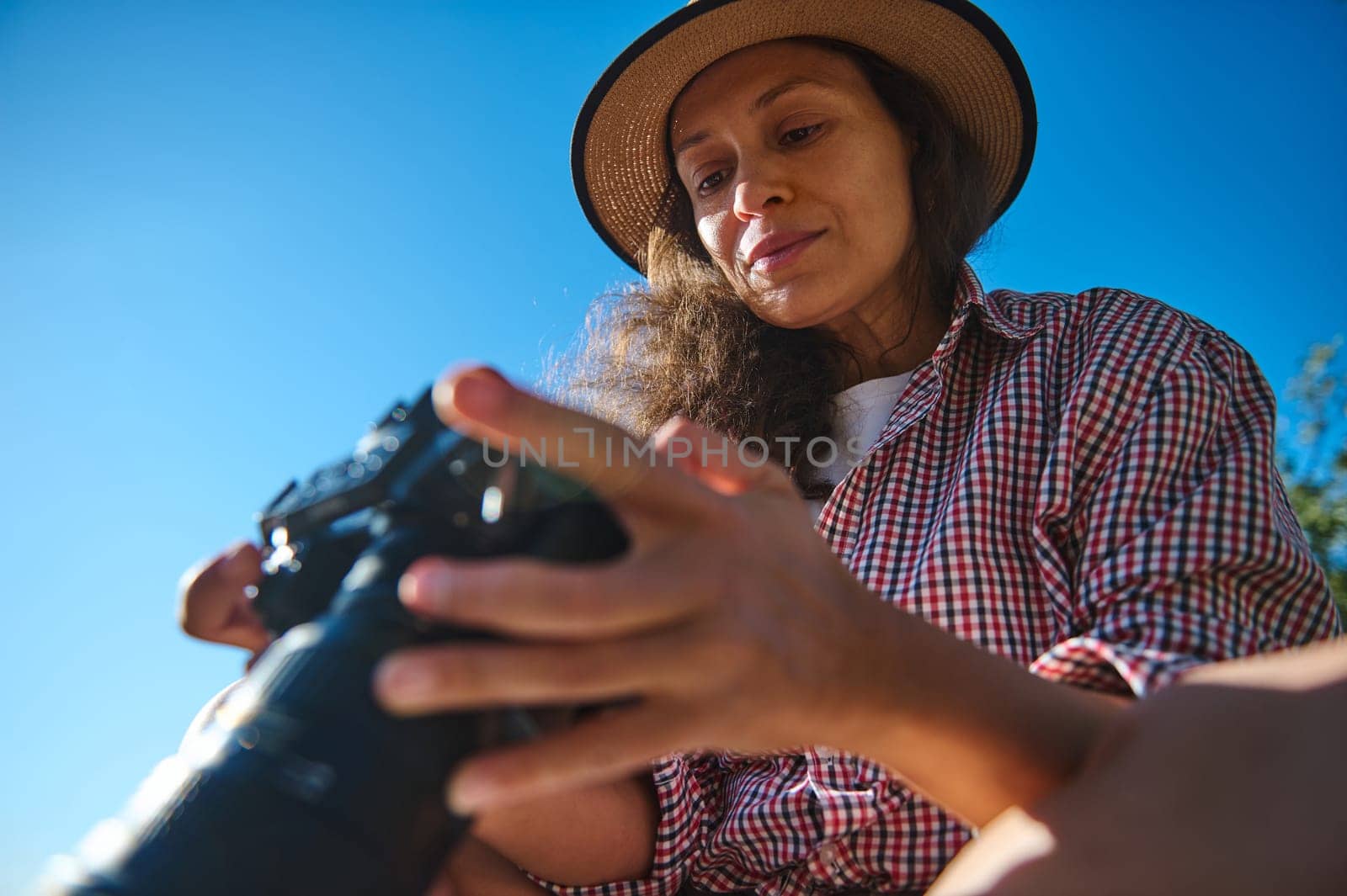 A woman wearing a straw hat and a plaid shirt examines her camera outdoors. The blue sky and natural light create a vibrant setting, symbolizing adventure and creativity.