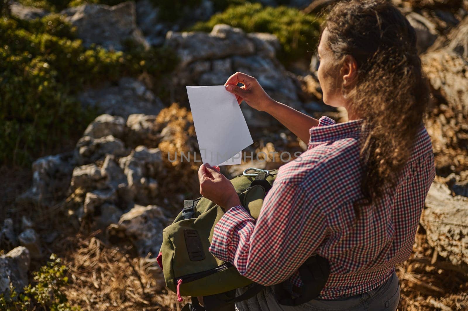 A woman in a plaid shirt examines a piece of paper while hiking through a rugged, rocky terrain. The scene conveys a sense of exploration and discovery amidst an adventurous outdoor environment.