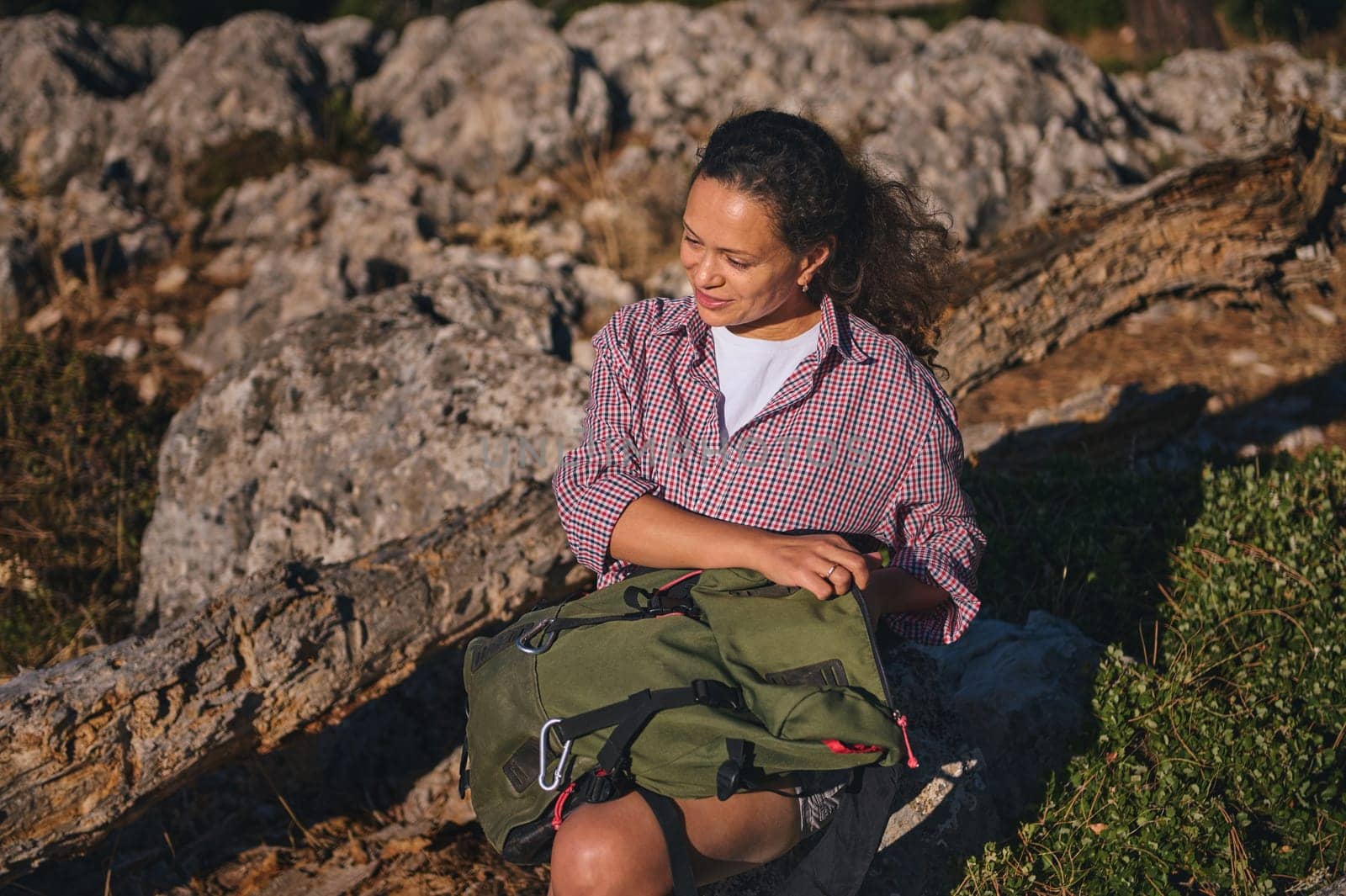 A woman relaxes on a rocky outdoor landscape, enjoying the sunlight and nature. She sits with a green backpack, wearing a casual checkered shirt. Sunlight enhances the peaceful mood.