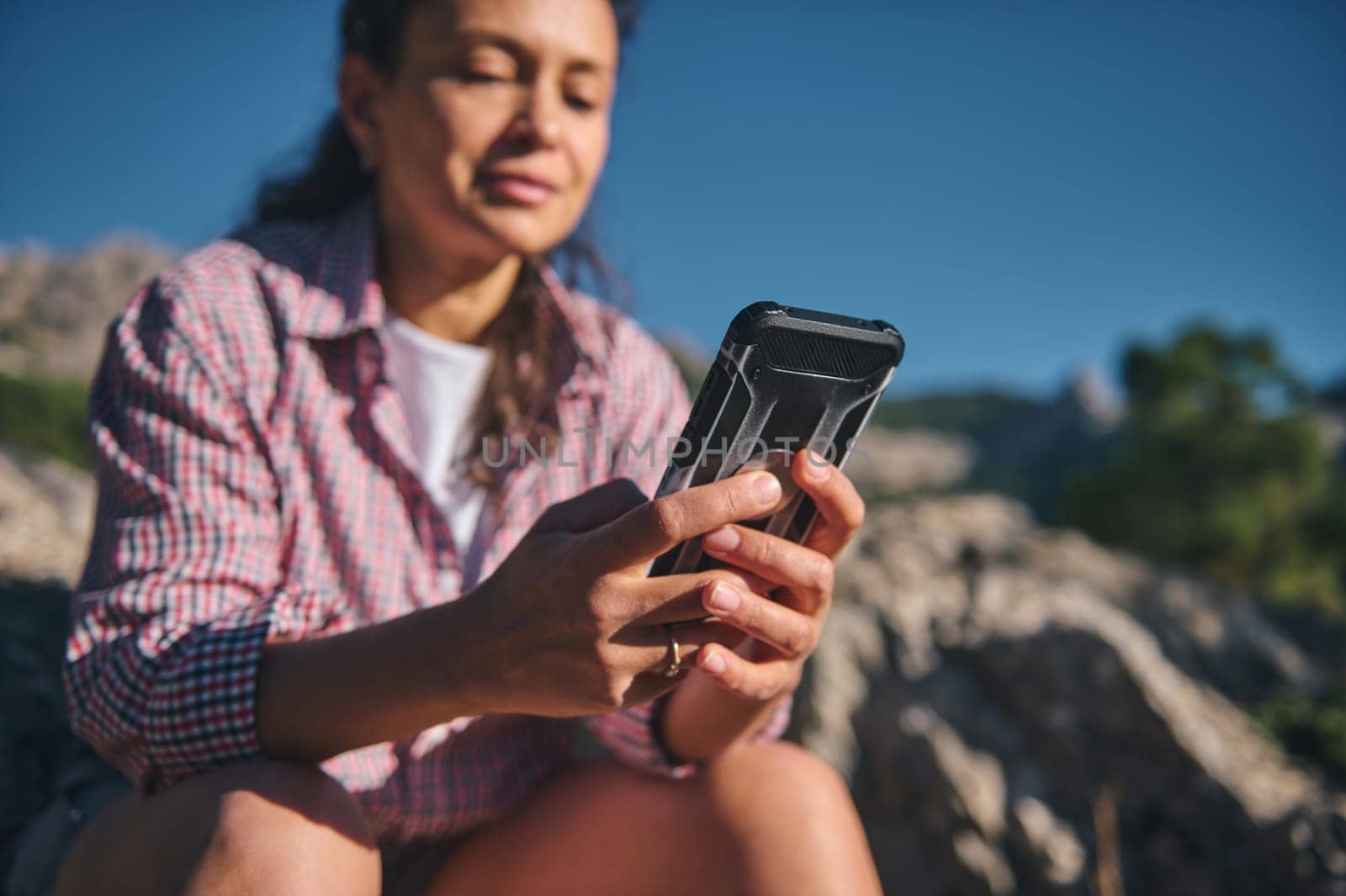 A woman enjoying a sunny mountain hike, using her smartphone while taking a break. The setting is tranquil and scenic, embodying relaxation, technology, and nature.