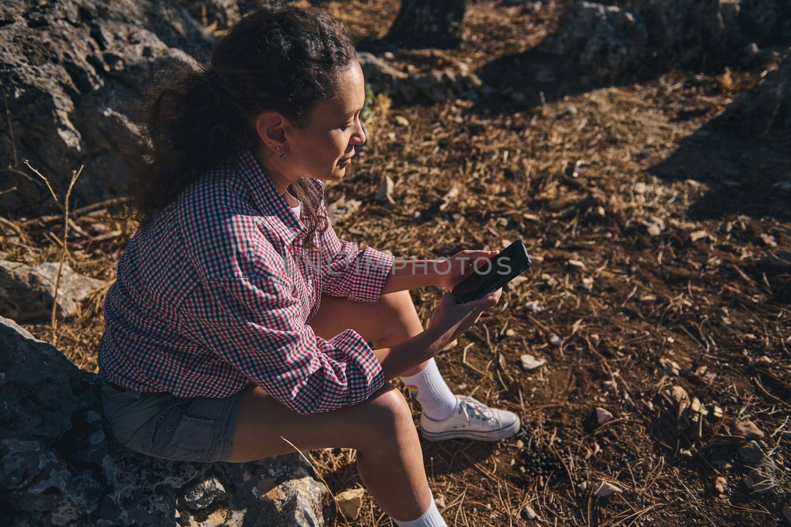 A young woman sits on rocks outdoors, engrossed in her smartphone. She enjoys the sunlight, dressed casually in a checkered shirt and shorts, surrounded by nature's tranquility.