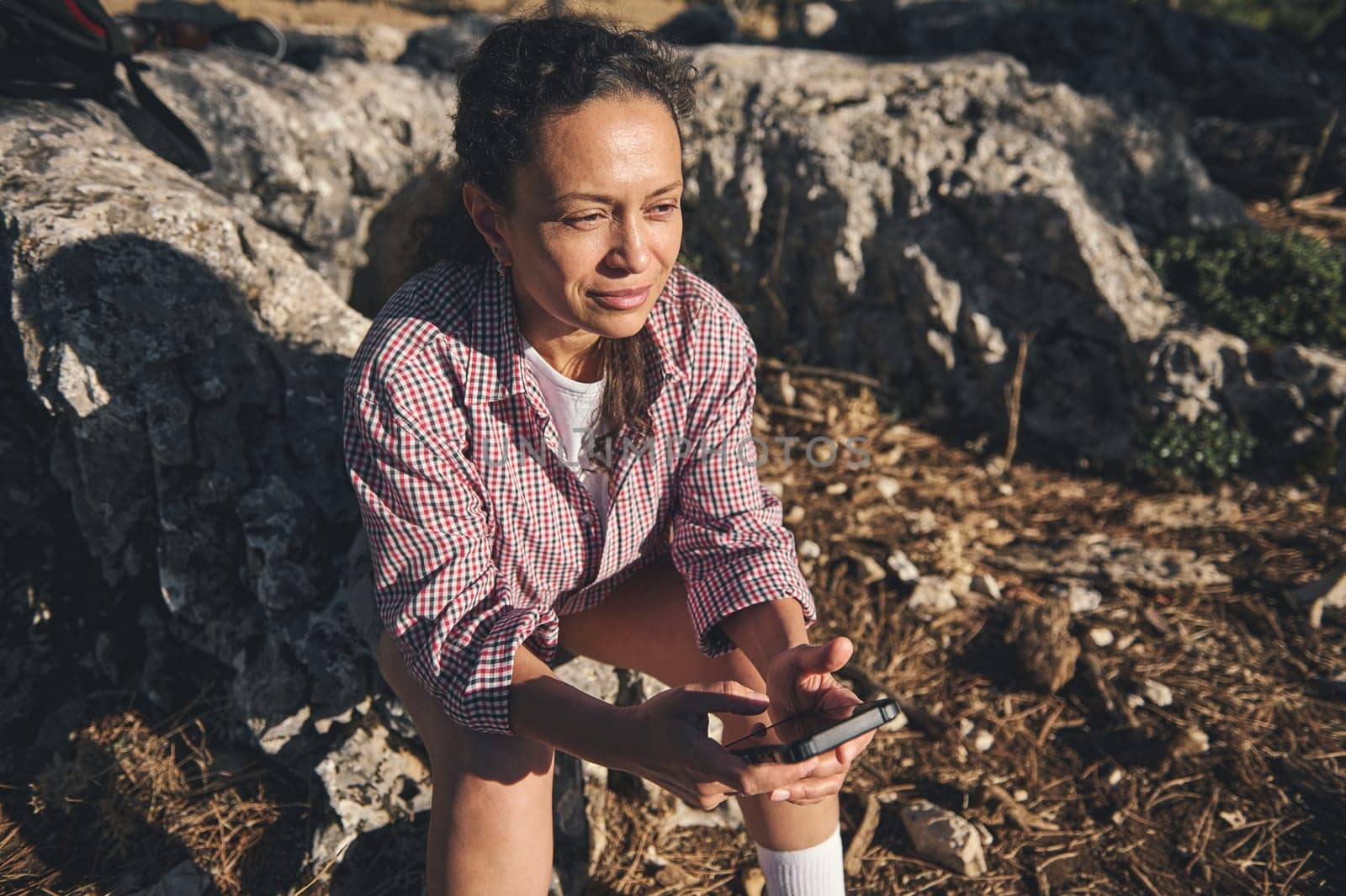A woman in a checkered shirt sits on rocks outdoors, holding a phone. She appears relaxed and thoughtful, basking in warm sunlight.