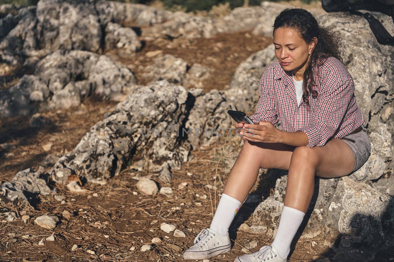 A woman in casual attire sits on rocks while using her phone in a natural setting. The scene suggests relaxation and a connection to nature, evoking a peaceful mood.