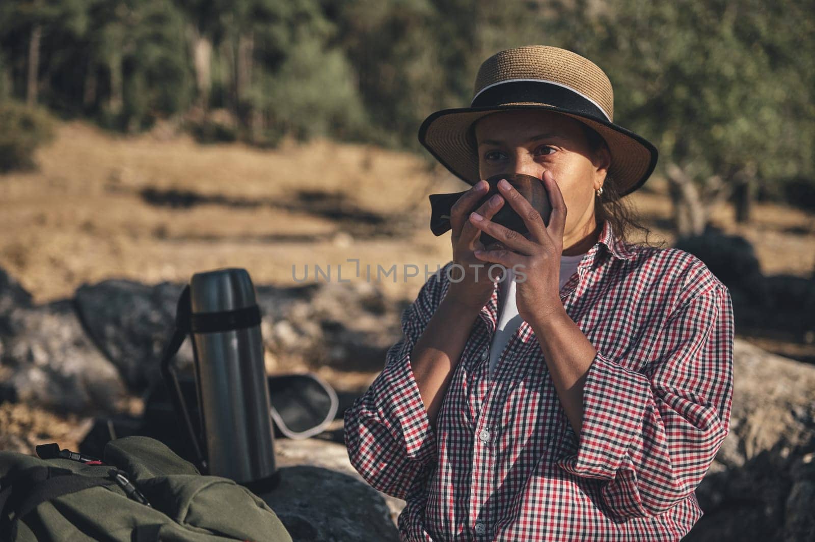 A woman in a checkered shirt and straw hat enjoys a hot drink while hiking. Surrounded by nature, she relaxes in the outdoors with a thermos nearby, capturing the essence of adventure and tranquility.