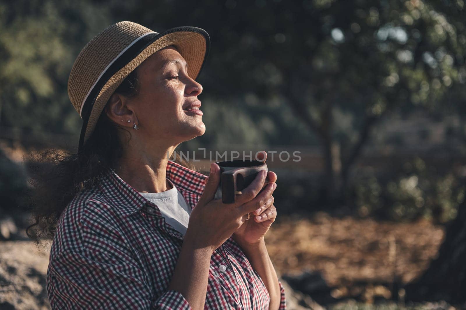 A woman in a straw hat savors a quiet morning outdoors, holding a cup of coffee. The serene expression suggests relaxation and contentment amidst nature, embracing tranquility.