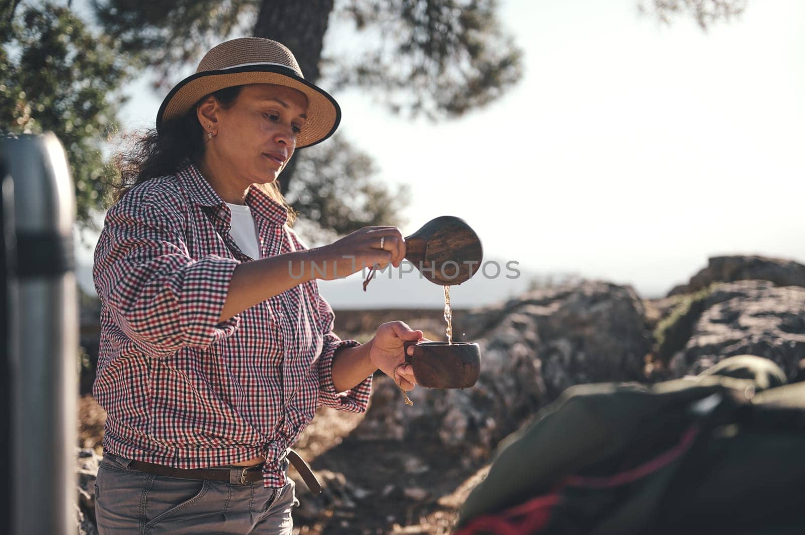 A woman in a hat pours coffee outdoors, surrounded by nature. Enjoying a relaxing moment, she is captured in a serene and tranquil setting, showcasing leisure and adventure.