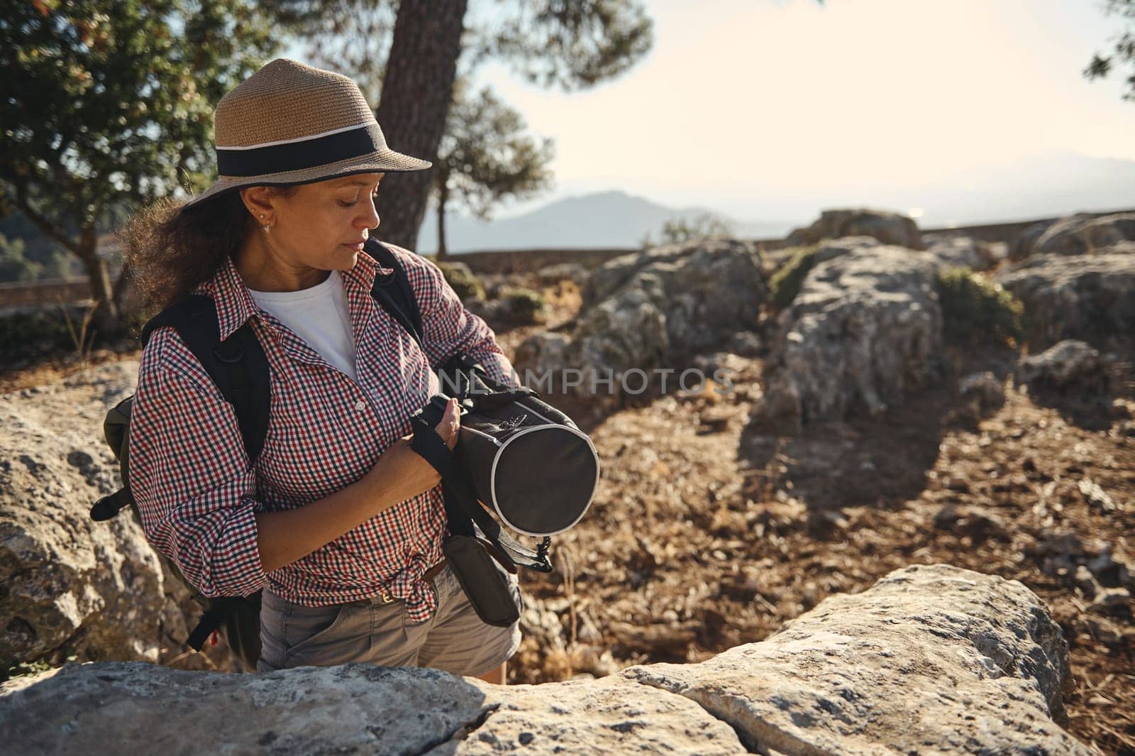 A woman wearing a hat explores a rocky landscape, carrying a backpack and camera. She enjoys nature and adventure under the clear sky, capturing moments while hiking in the outdoors.