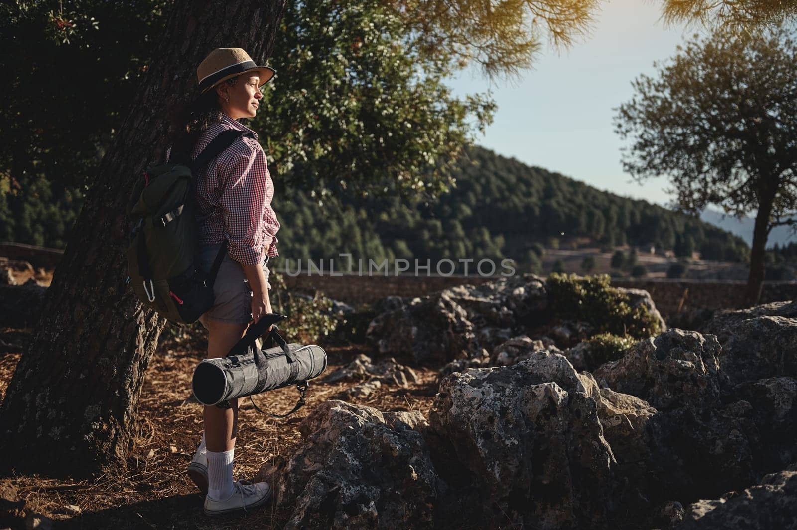 A woman rests under a tree with a backpack and yoga mat in a serene outdoor setting. She is surrounded by rocky terrain and lush forests under a sunny sky, conveying adventure and tranquility.