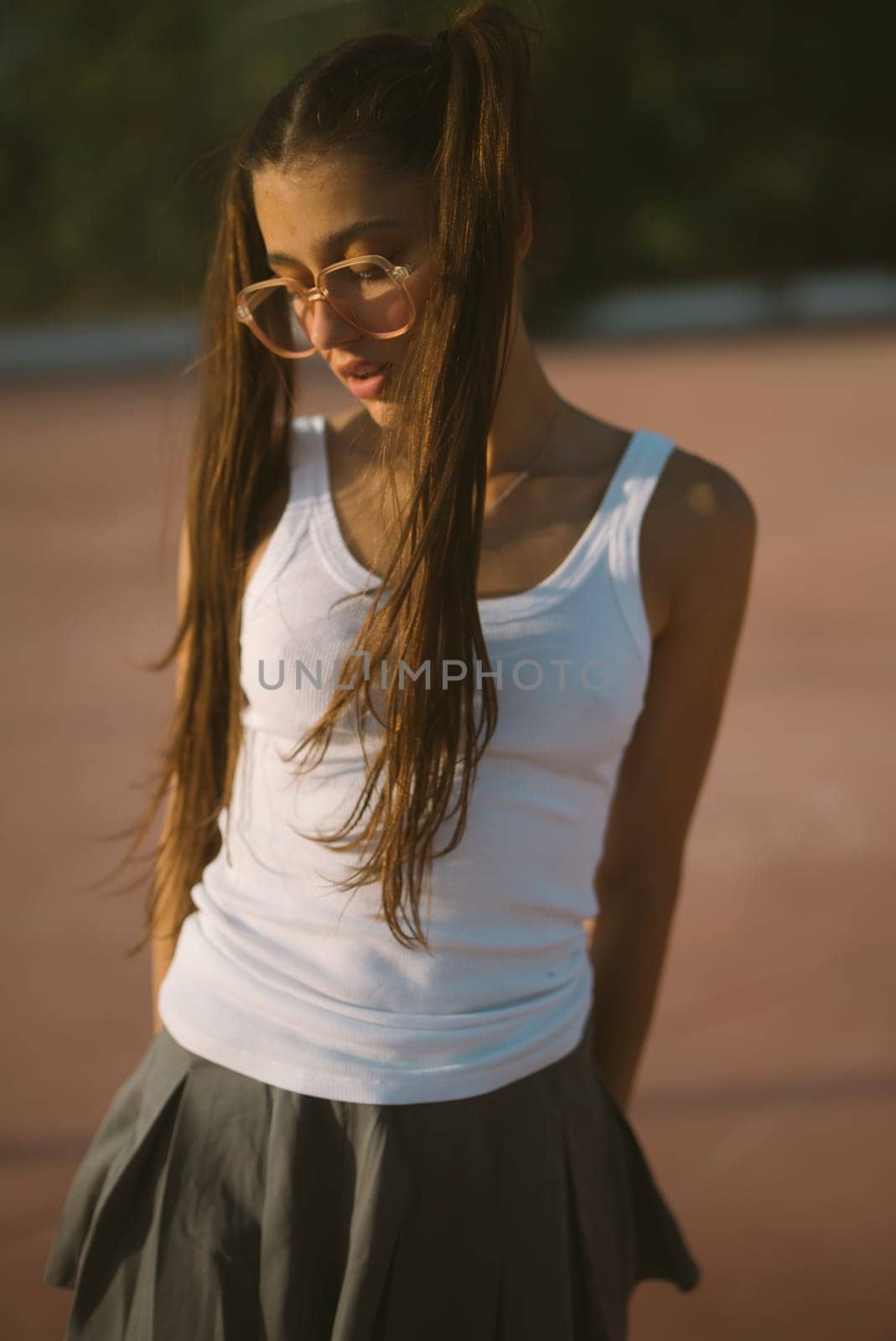 A playful young girl with long hair, wearing a white tank top and a skirt, exudes confidence and charm.