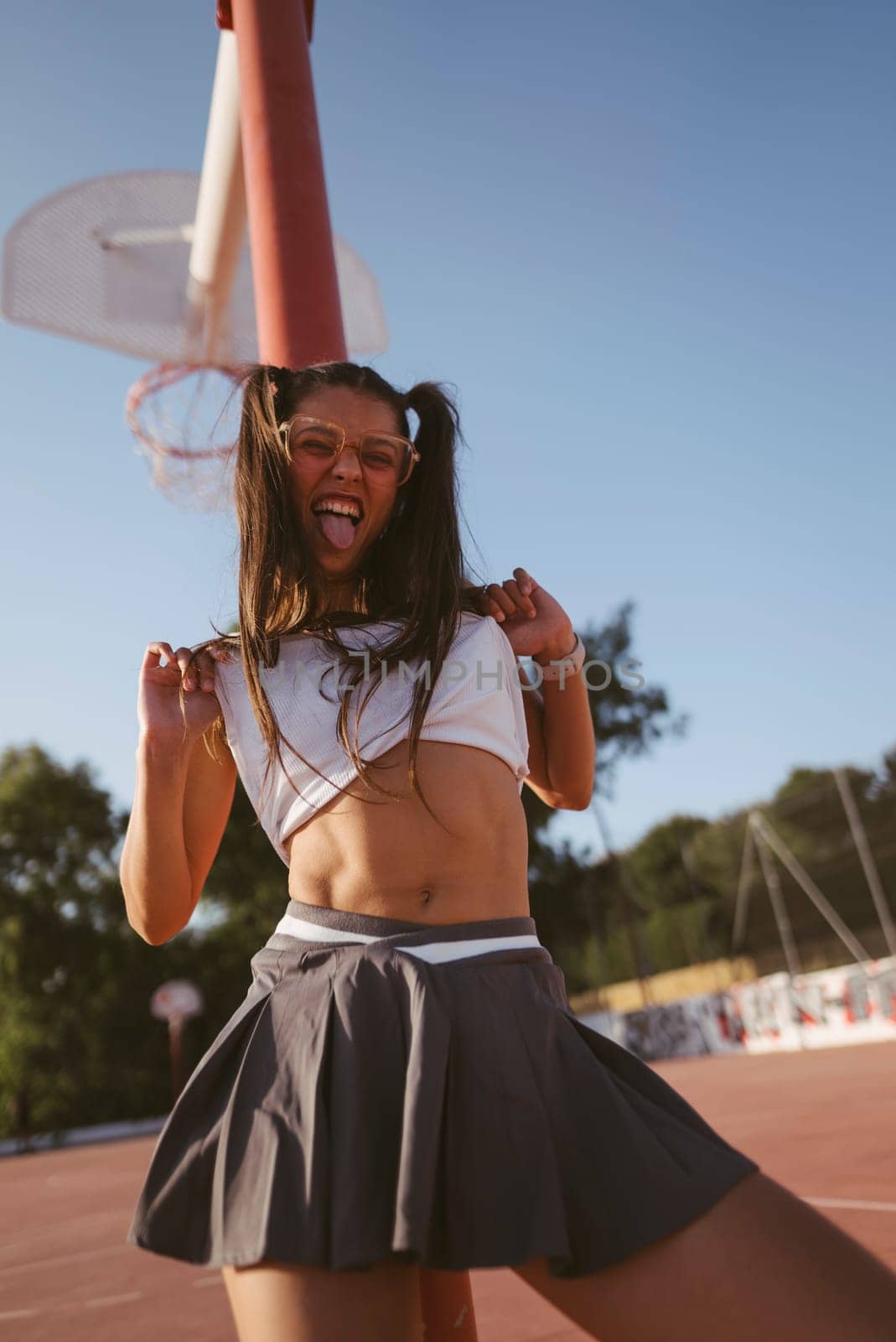A cheerful young woman in a lively cheerleader outfit poses joyfully outdoors on a sunny basketball court