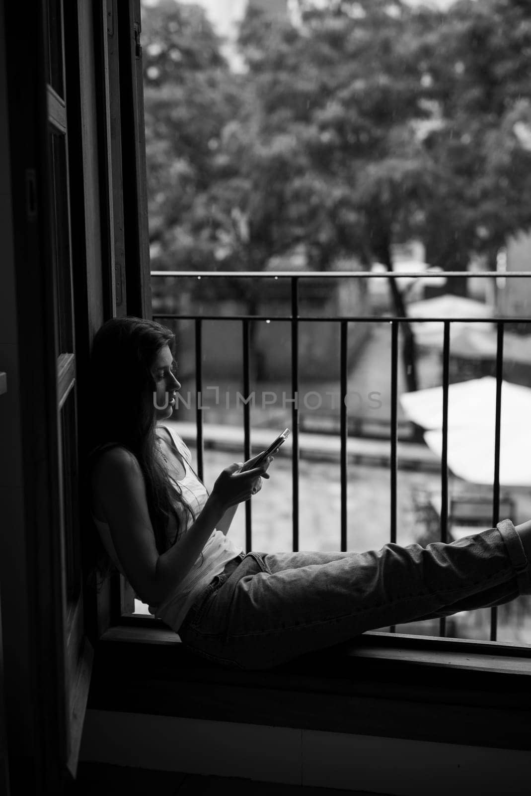 A young woman sits by the window, deeply absorbed in her phone, capturing a fleeting moment of solitude and calmness