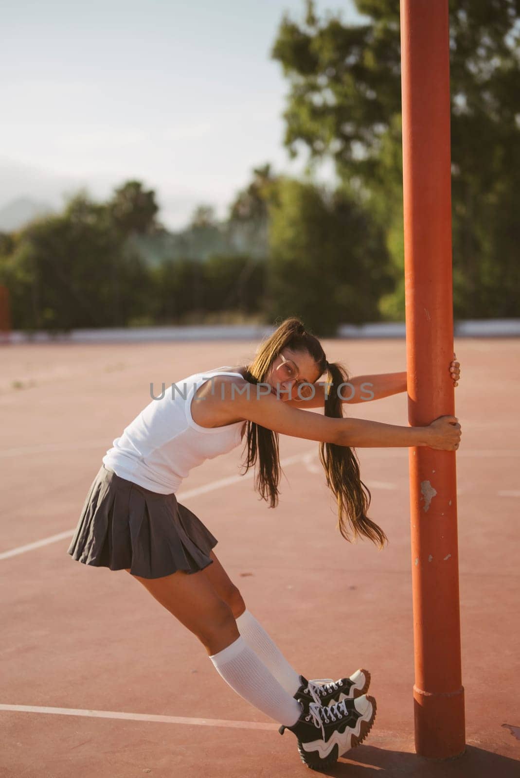 A young woman dressed in a sporty outfit is stretching gracefully by a pole on a bright sunlit outdoor court