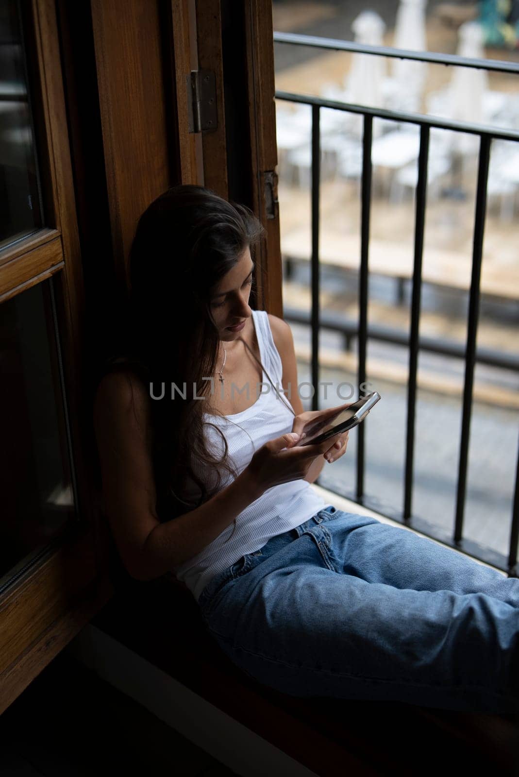 A woman in casual wear is seated by the window, engaged with her smartphone in a cozy, relaxing atmosphere