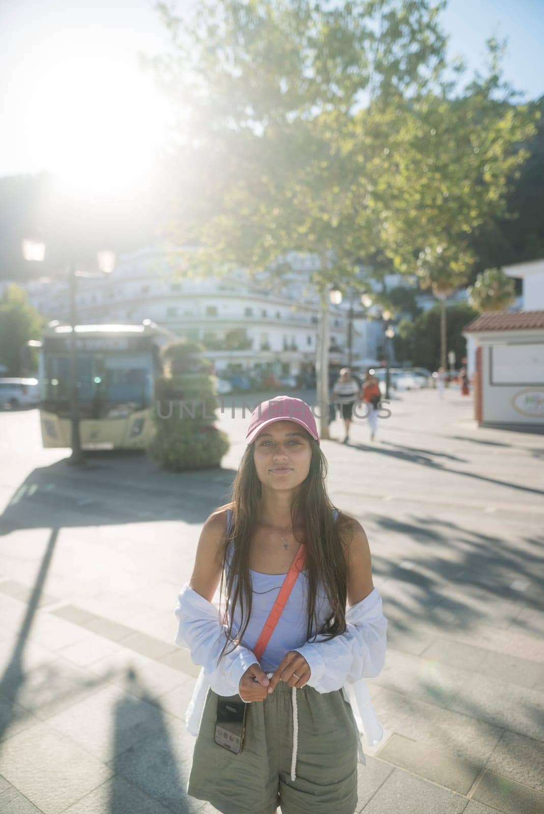 In a vibrant urban environment illuminated by bright sunlight, a stylish young woman poses joyfully outdoors