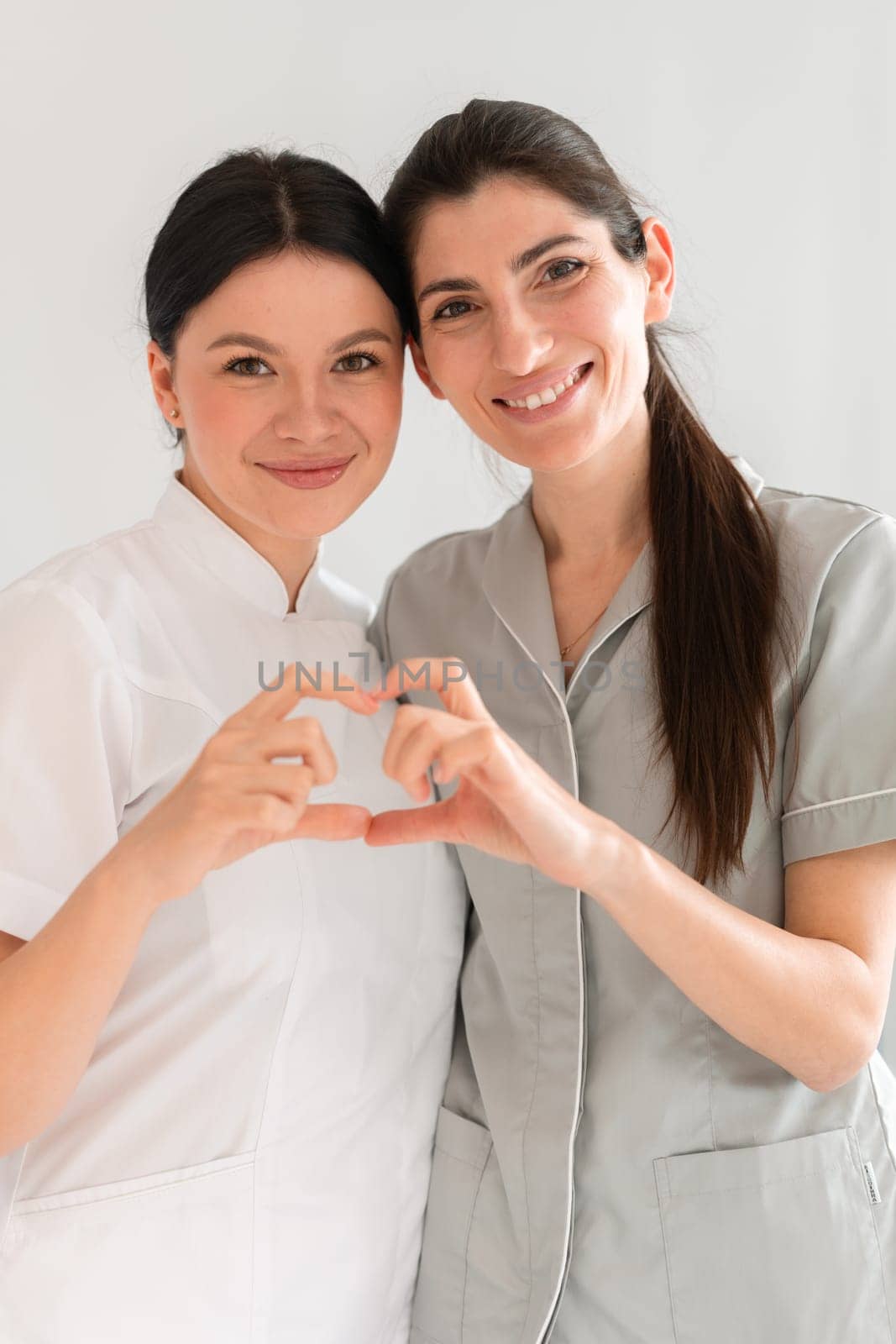 Happy female doctors making heart shape with hands at clinic. Smiling medical professionals are looking at camera while standing together in hospital.