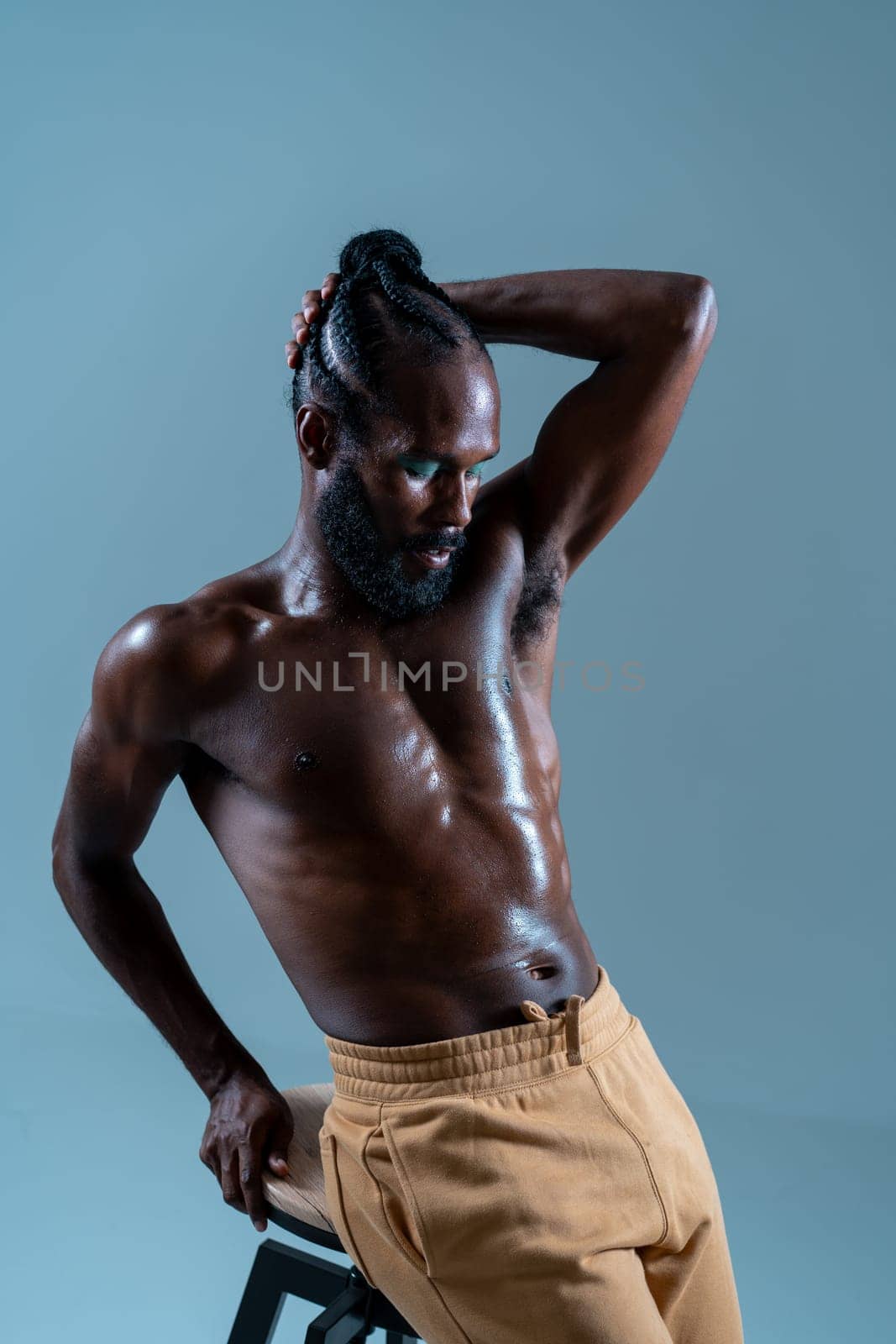 African American shirtless gay model posing while leaning on stool over blue background. Muscular homosexual man is with hand behind head in studio.