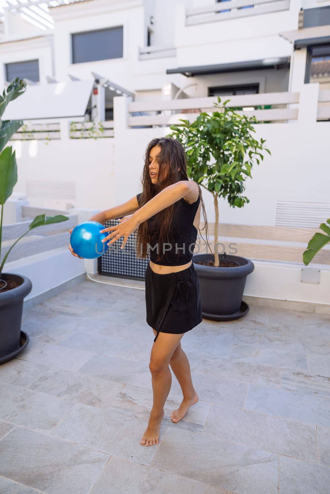 A cheerful young woman is playfully engaging with a vibrant blue ball in a sunny outdoor setting filled with greenery