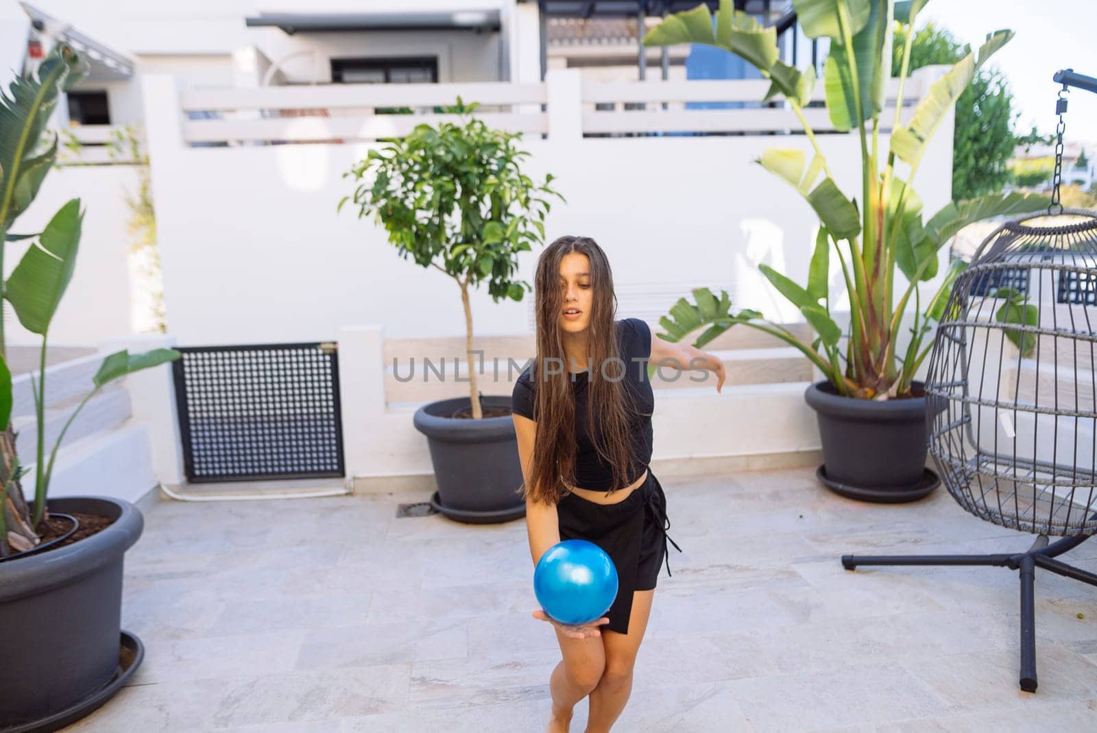 A young woman gracefully throws a vibrant blue ball in a stylish outdoor area that is surrounded by lush plants