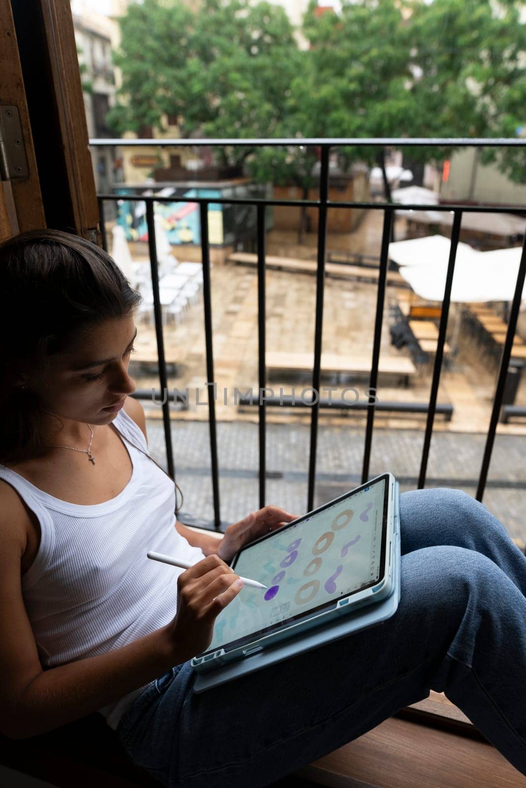 A young woman sits comfortably on her balcony, sketching creative ideas on her tablet while relaxing outside