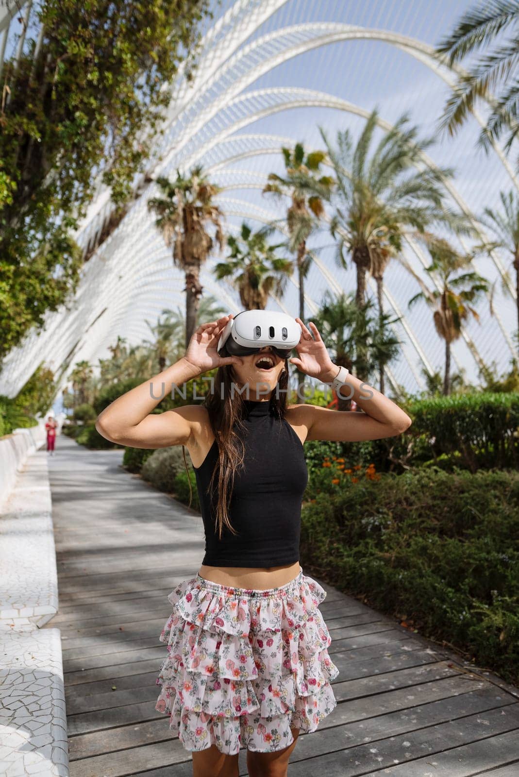 A stylish young woman is engaged with a virtual reality headset while surrounded by palm trees and modern architecture