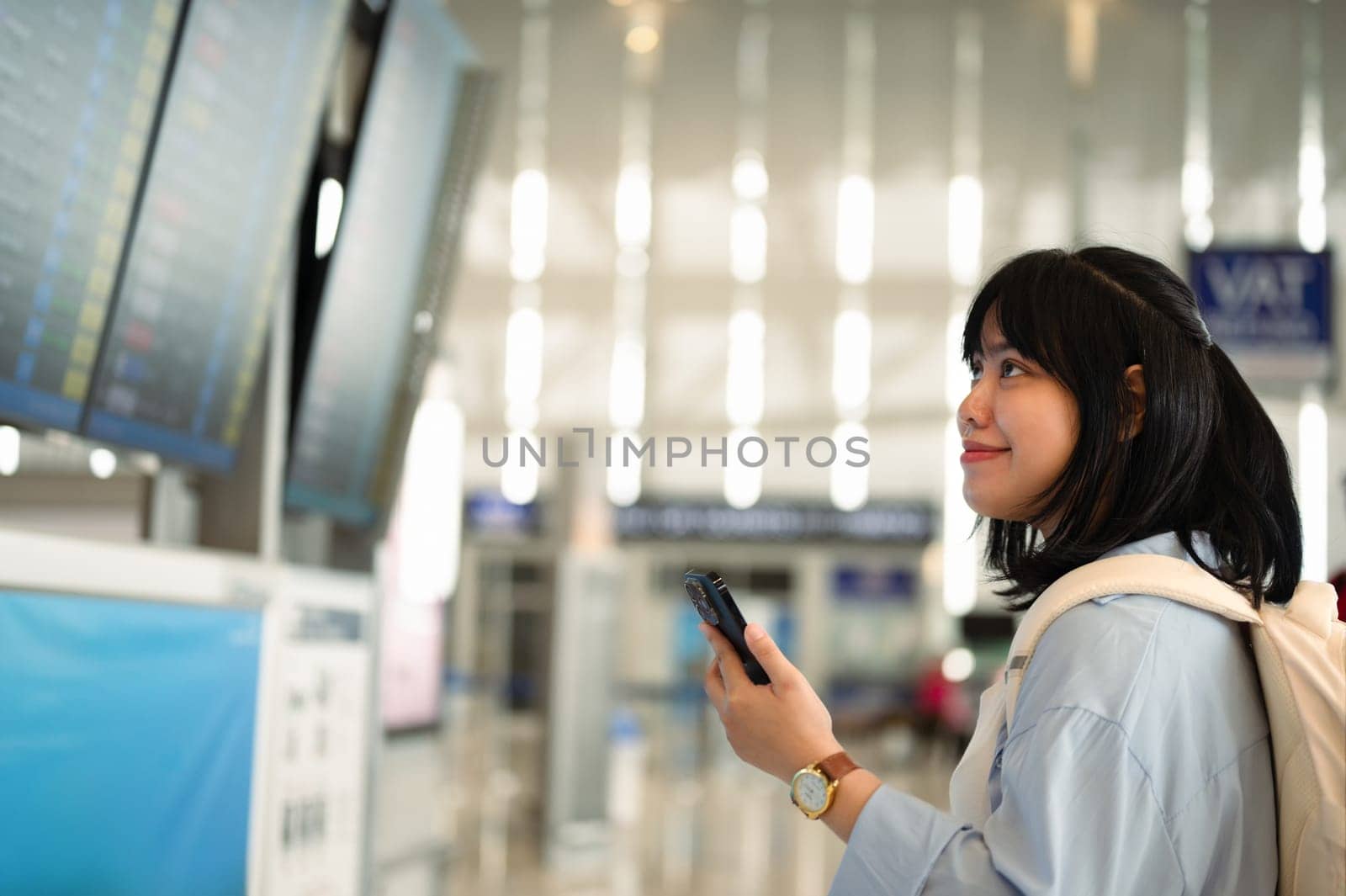 Smiling young woman holding mobile phone and checking her flight schedule at airport.