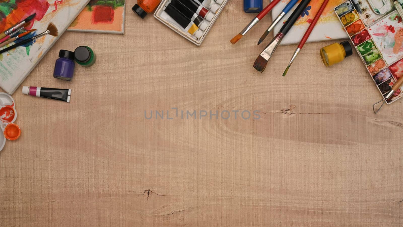 A wooden table with a variety of art supplies including paint brushes, watercolor and canvas.