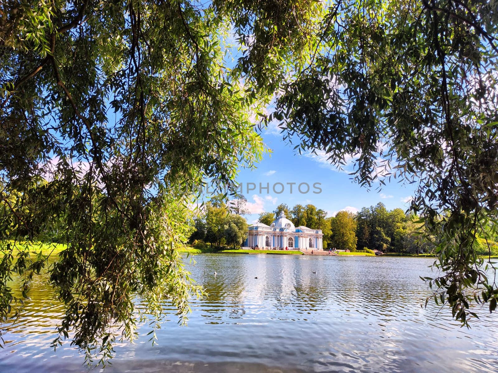 A historic building stands beside a calm lake, framed by vibrant foliage under a clear blue sky, showcasing peaceful nature.