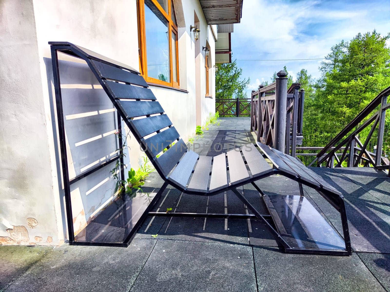 A sleek lounge chair made of black slats rests on a terrace, enhanced by natural light and lush green surroundings.