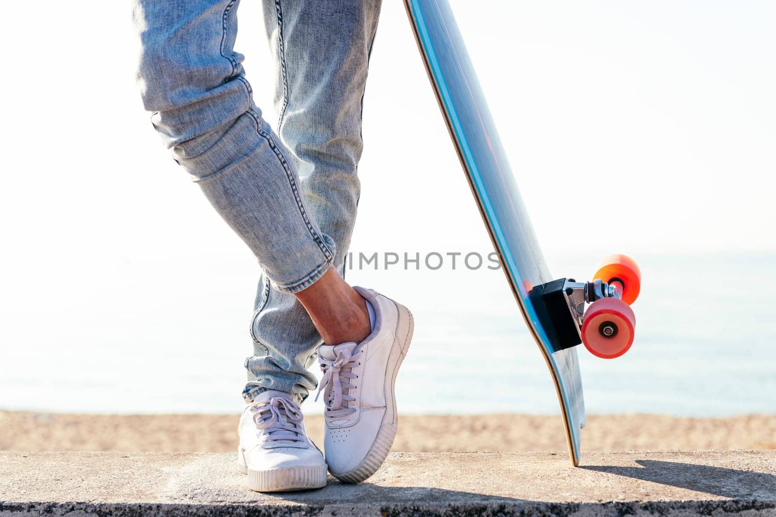unrecognizable stylish skater in jeans and sneakers standing next to skateboard on street on sunny day in summer