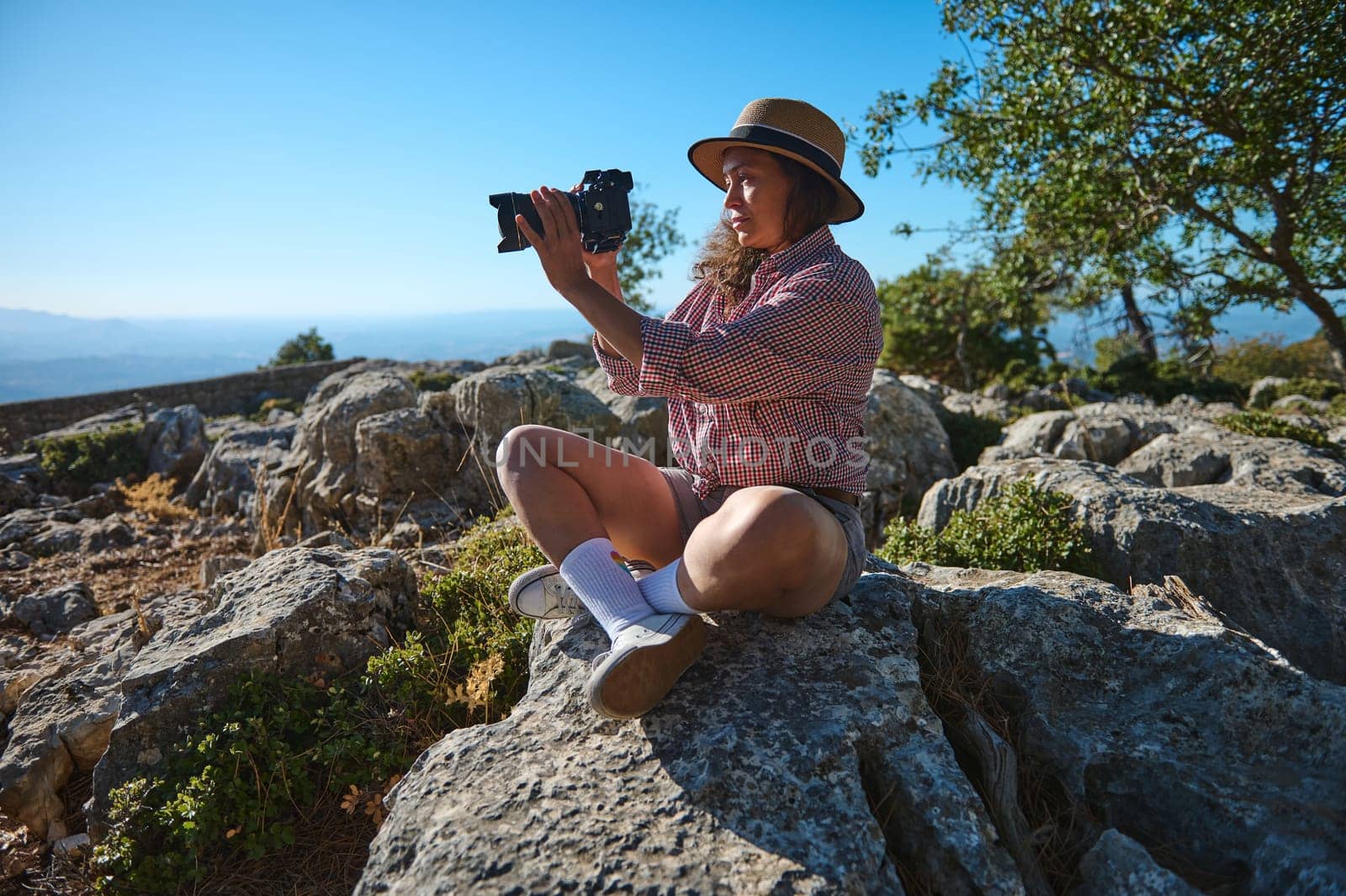 A woman in casual attire is sitting on rocks, capturing the scenic landscape with a camera under a clear blue sky.