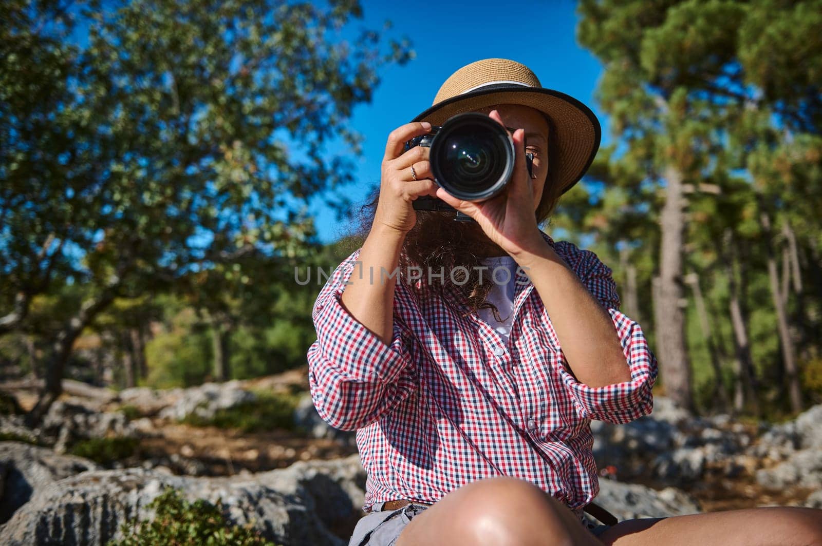 A woman wearing a hat holds a camera, capturing moments in a lush forest. The bright day highlights her passion for photography amidst nature. An image of exploration and creativity.