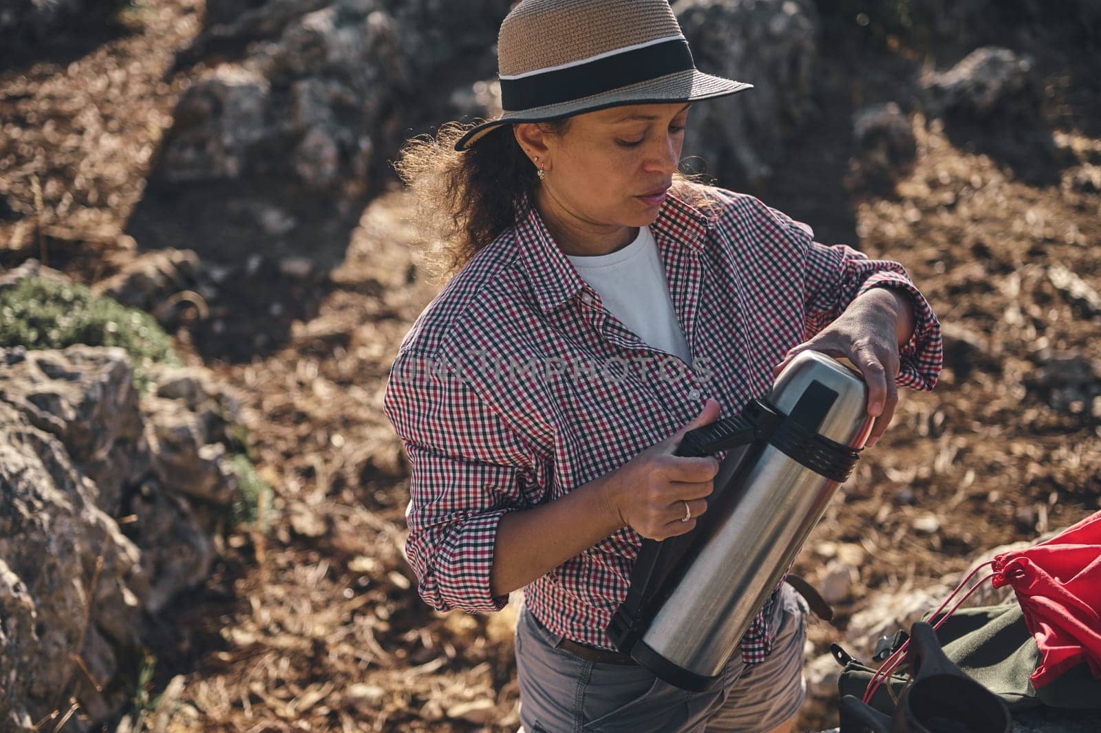 A woman in a checked shirt and hat pours a hot drink from a thermos during a hiking adventure. She seems relaxed and content, embracing the tranquility of nature.