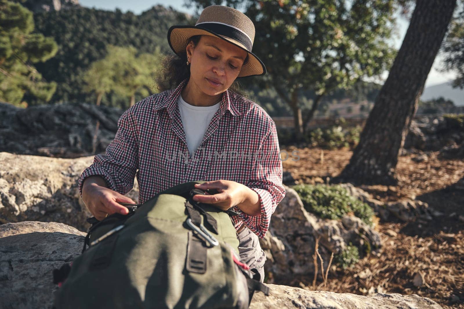A woman in a sun hat and checked shirt sits outdoors with a backpack. She enjoys the tranquil natural surroundings, soaking in the serene atmosphere of the forest setting.