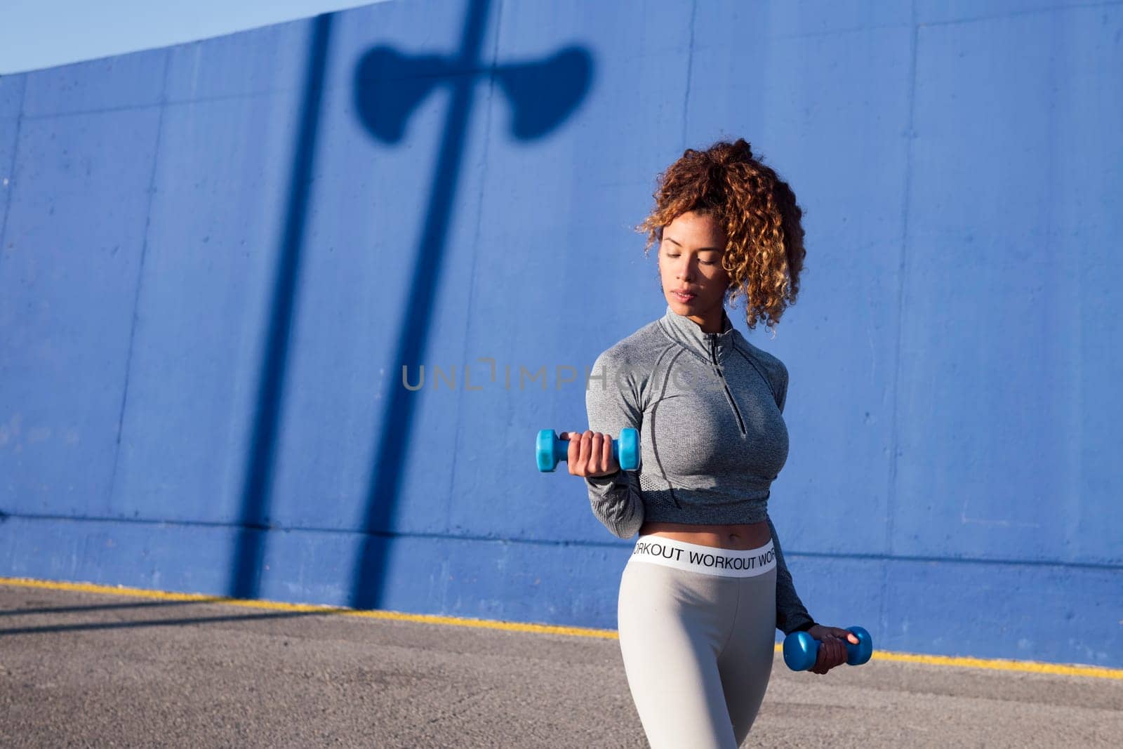 latin female athlete in sports clothes working out with dumbbells on urban roadway against blue wall in sunlight, concept of urban sport and healthy lifestyle
