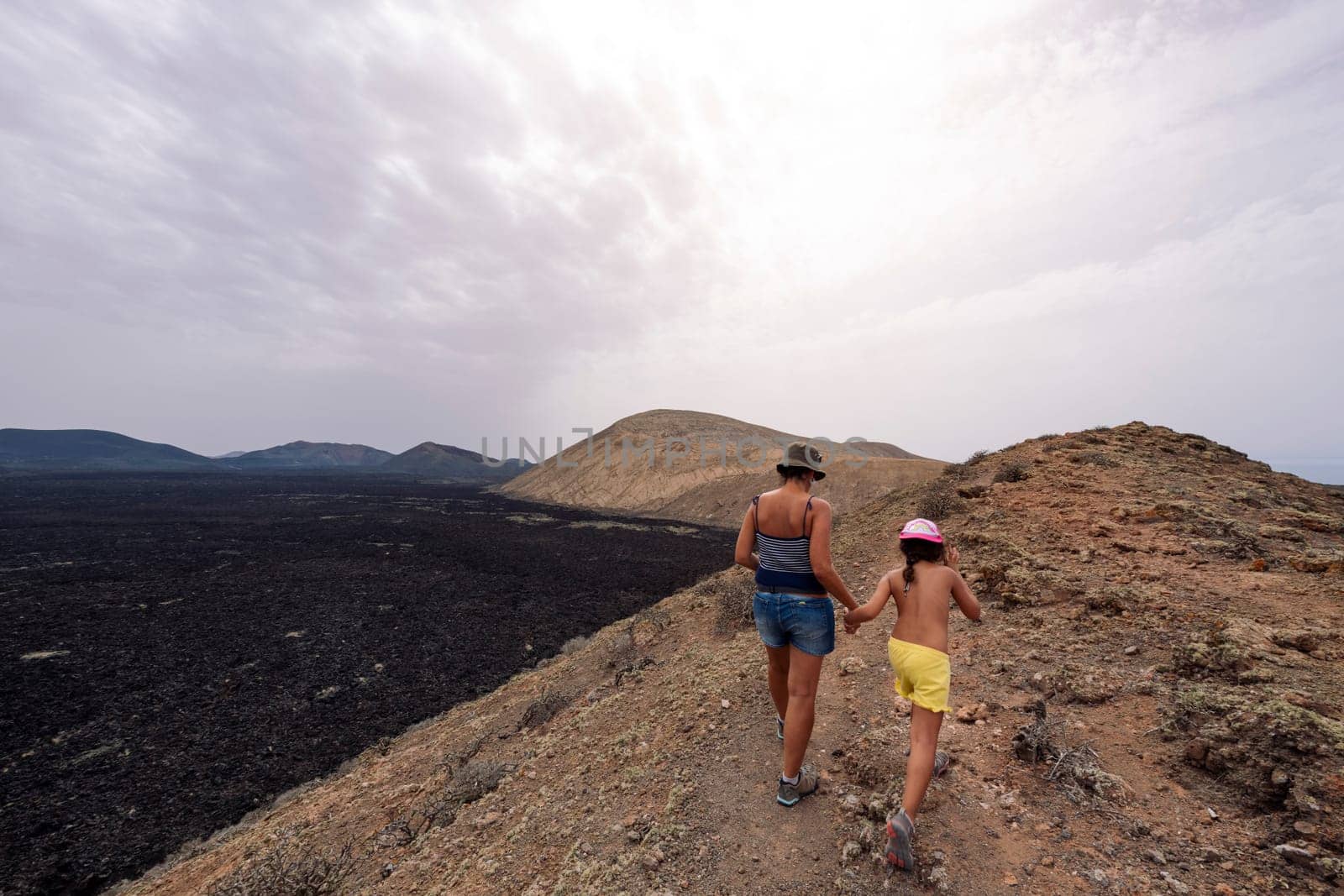 rear view of mom with girl strolling holding hands by rough trail in crater of volcano in Natural Park of Lanzarote in Canary Islands, Spain, concept of active tourism and adventure travel