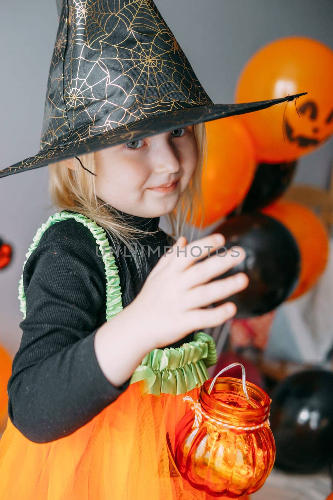 Children's Halloween - a girl in a witch hat and a carnival costume with airy orange and black balloons at home. Ready to celebrate Halloween.