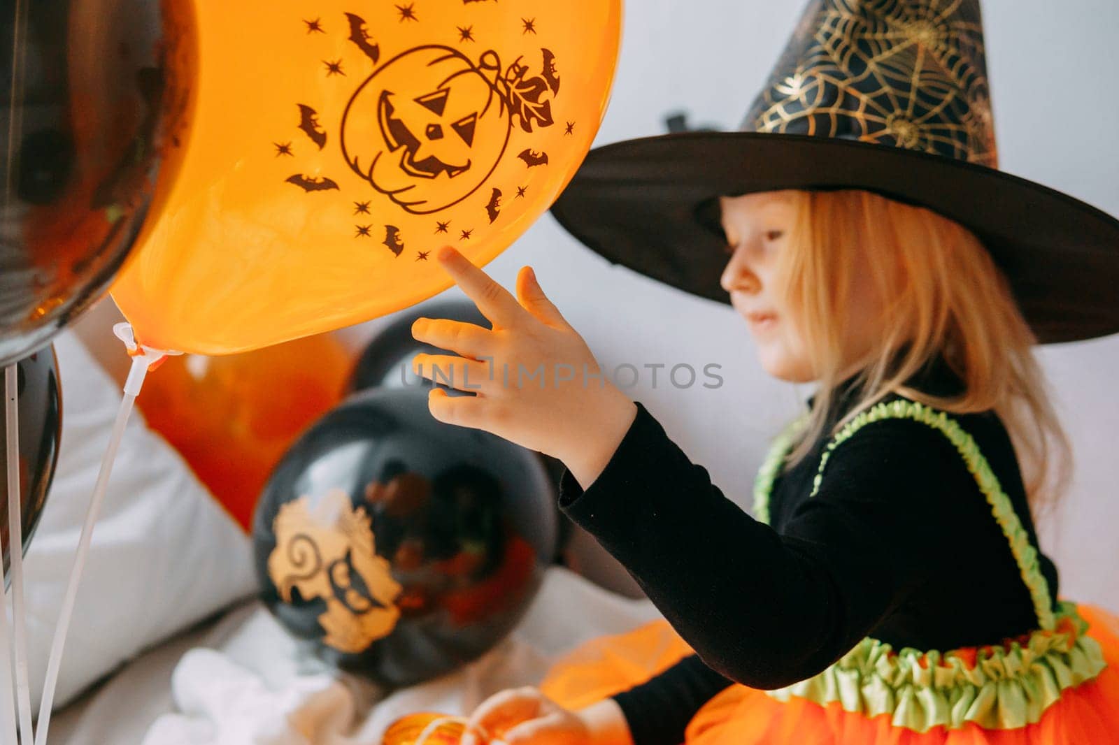 Children's Halloween - a girl in a witch hat and a carnival costume with airy orange and black balloons at home. Ready to celebrate Halloween.