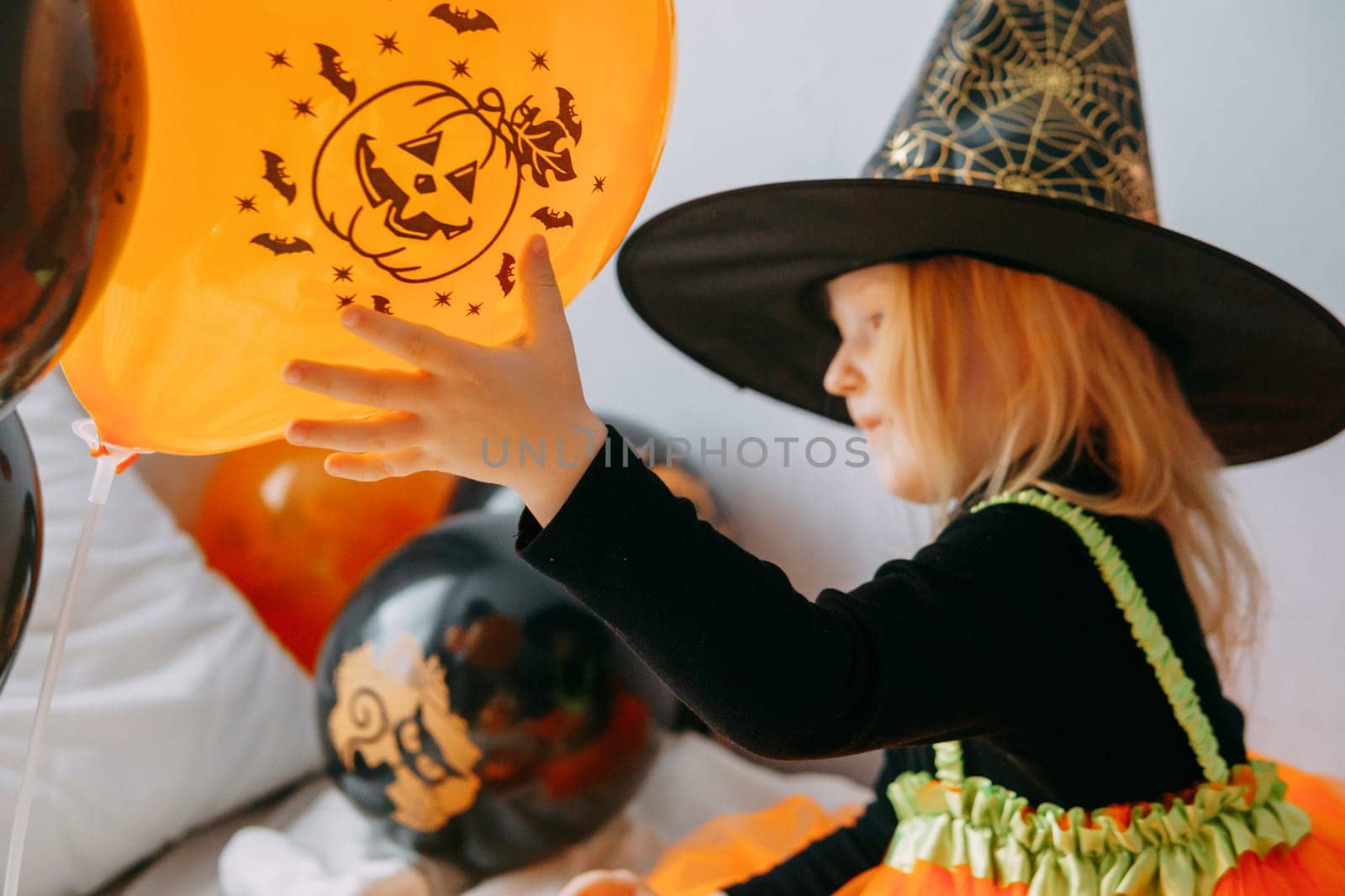 Children's Halloween - a girl in a witch hat and a carnival costume with airy orange and black balloons at home. Ready to celebrate Halloween.