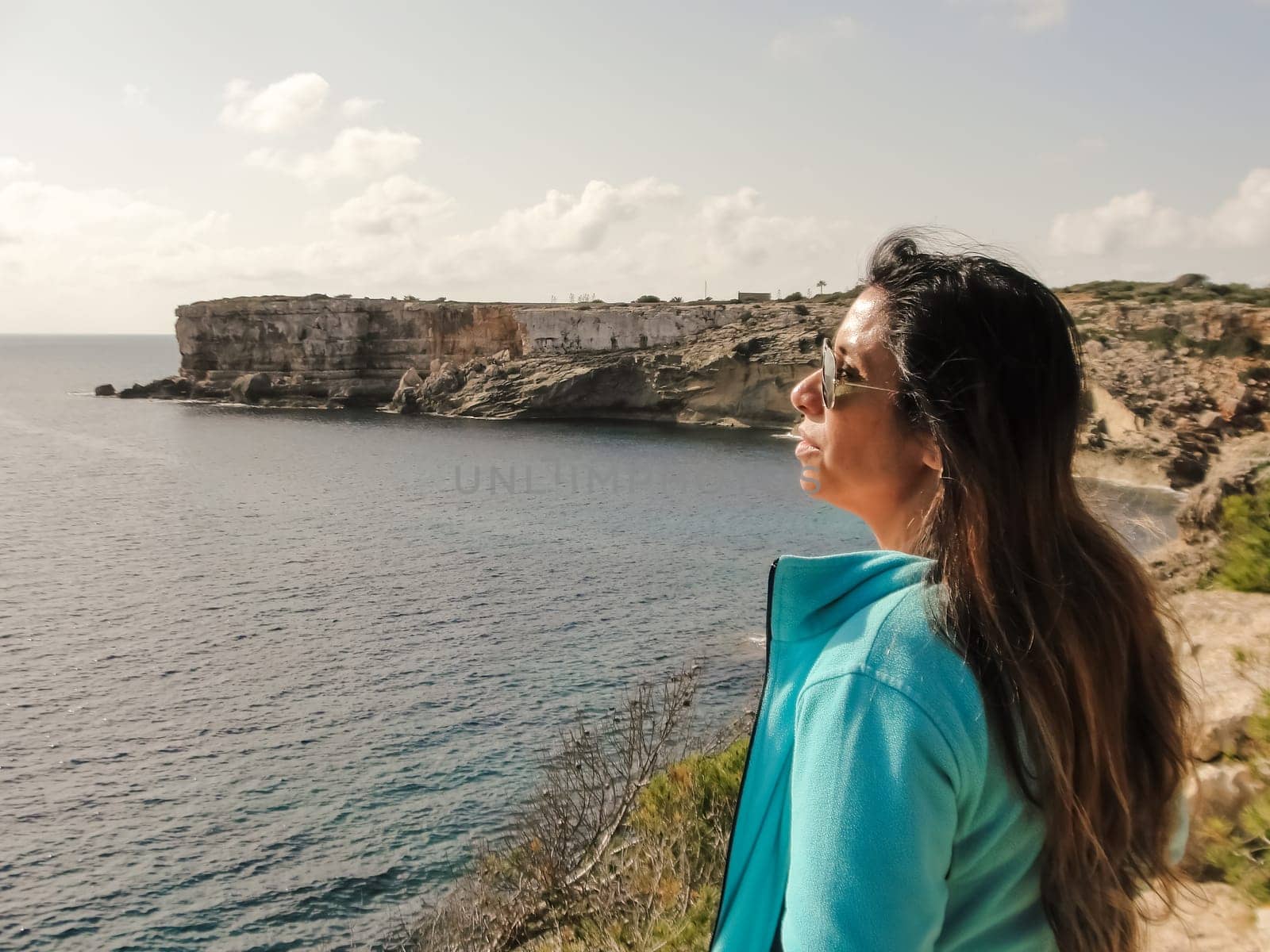 Woman with long hair gazes at ocean from a rocky cliff under sunny skies, latin woman looking out over the mediterranean sea cliffs, in mallorca balearic islands spain,