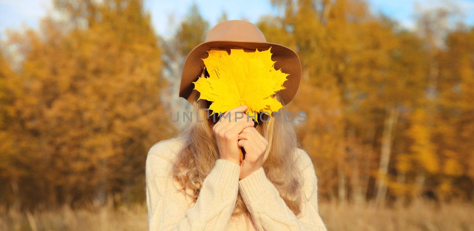 Portrait of happy cheerful woman covering her face with yellow maple leaves in autumn park, enjoying warm sunny weather, fall season, nature background