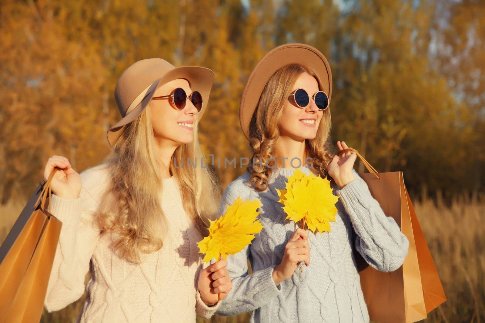 Happy young two women friends with autumn yellow leaves, holding shopping bags, stylish smiling girlfriends standing together outdoors, in sunny park, warm fall season