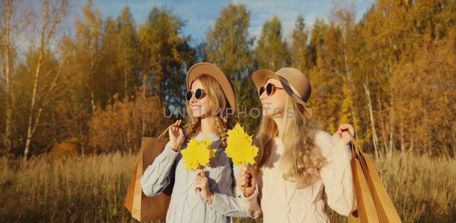 Happy young two women friends with autumn yellow leaves, holding shopping bags, stylish smiling girlfriends standing together outdoors, in sunny park, warm fall season