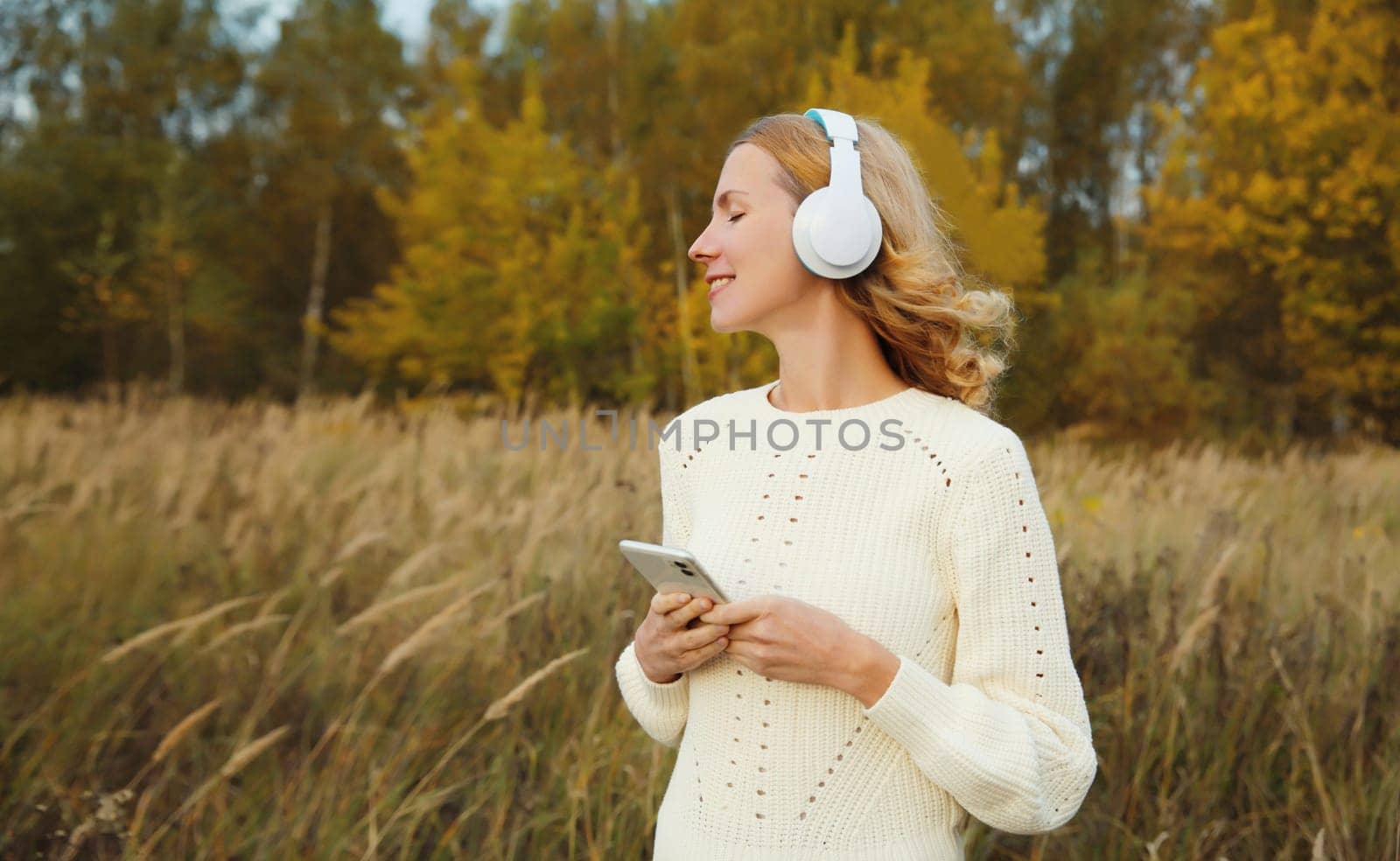 Happy inspired young woman enjoying listening to music in headphones with phone outdoors in autumn forest with yellow leaves