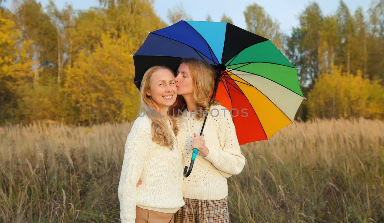 Adult daughter kissing her happy smiling mature mother holding colorful umbrella walking in autumn park with yellow leaves