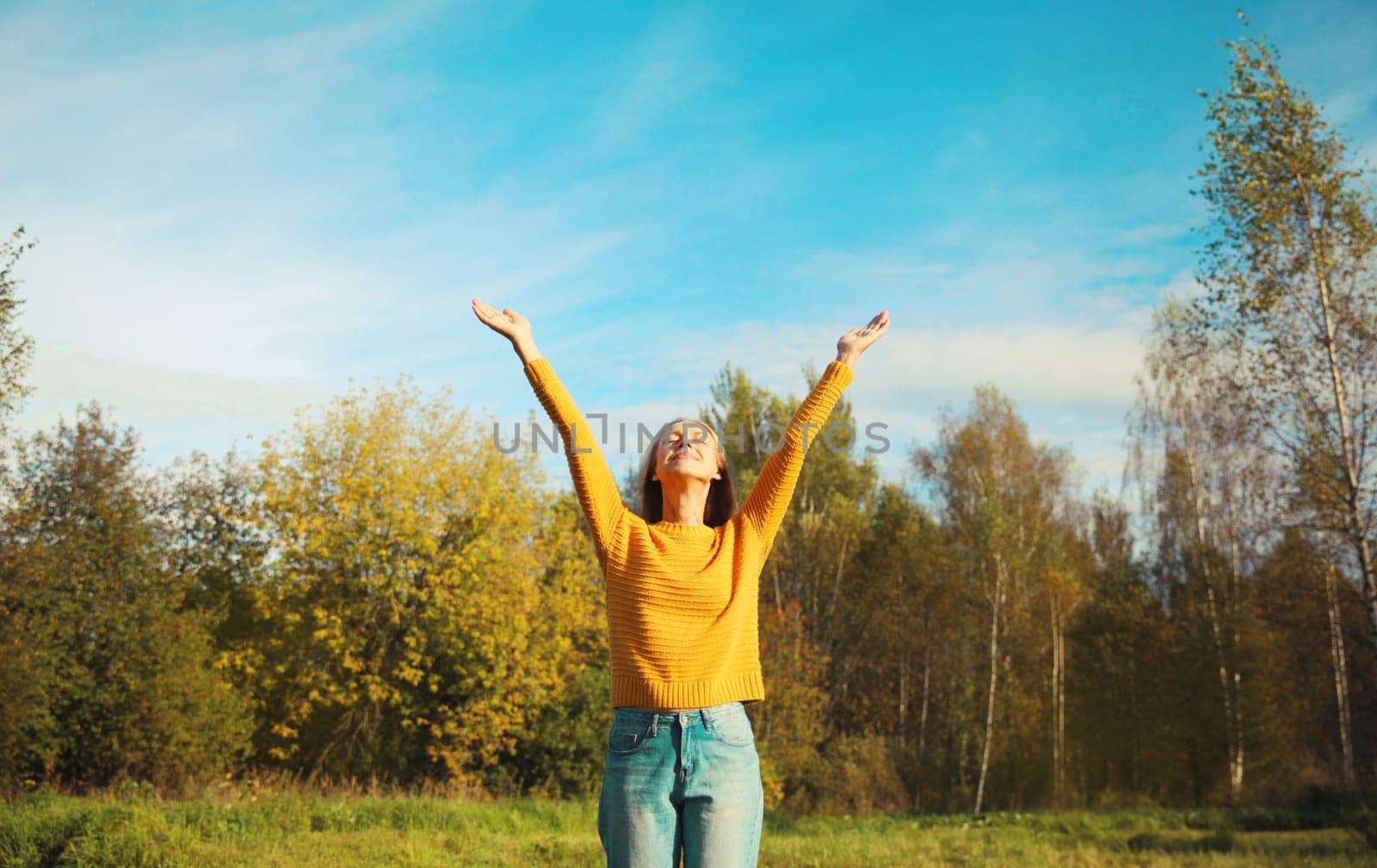 Happy smiling joyful woman raising her hands up in autumn park with yellow leaves, blue sky, enjoying warm sunny weather, fall season, nature background