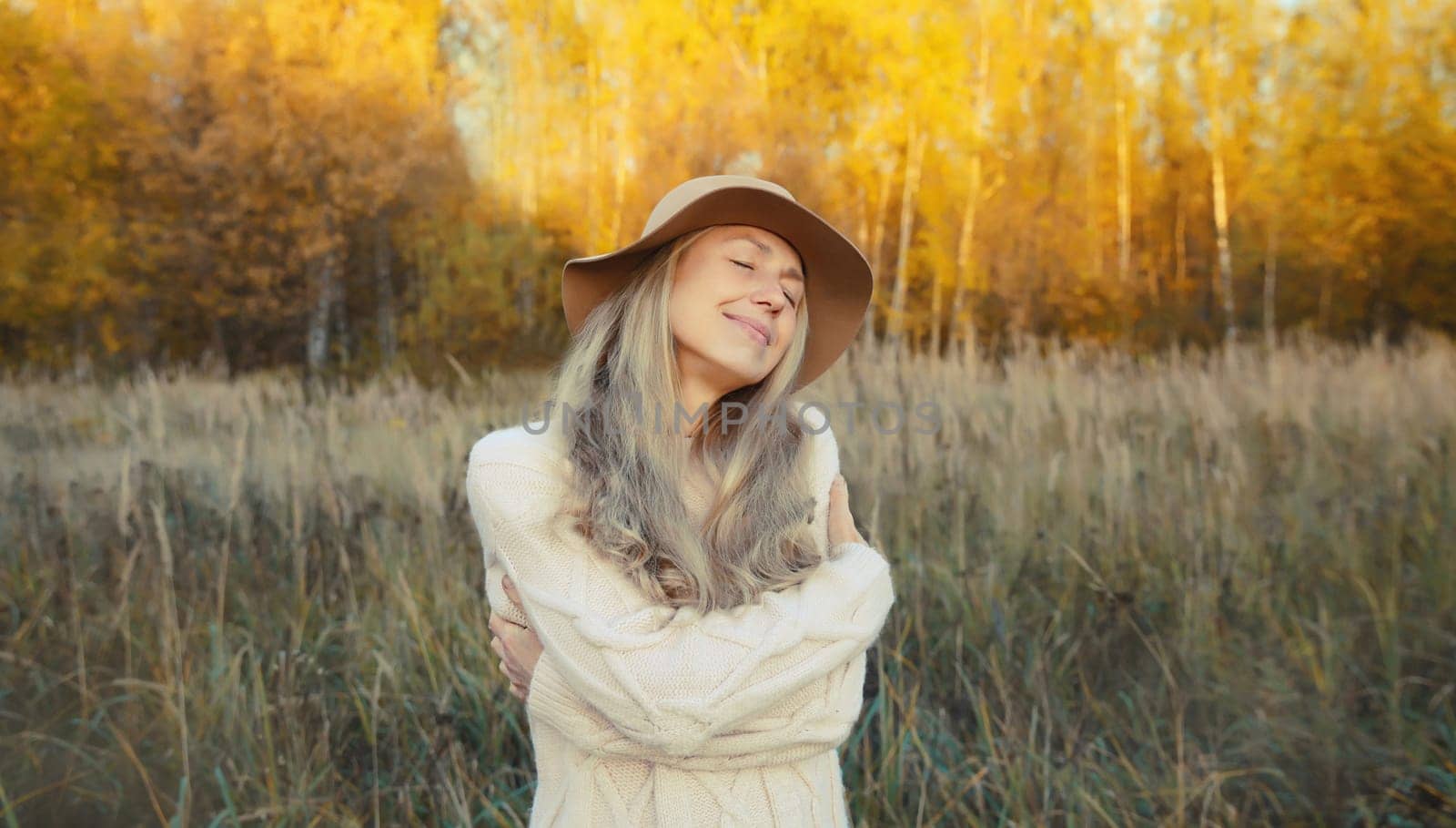 Happy smiling mature woman hugs herself in autumn park enjoying warm weather on sunny day outdoors.